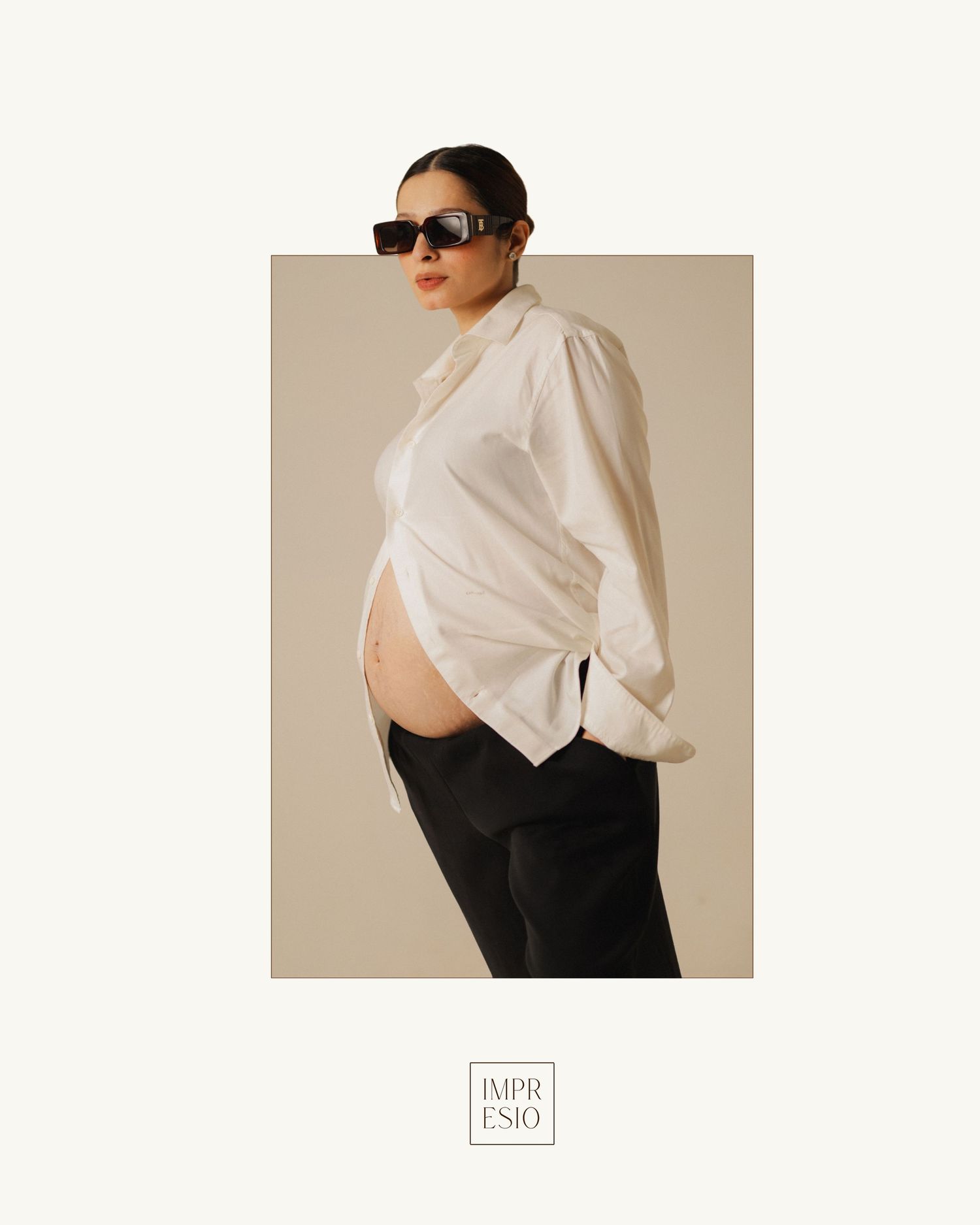 Pregnant woman in white oversized shirt, black pants, and sunglasses posing sideways for Impresio maternity fashion.