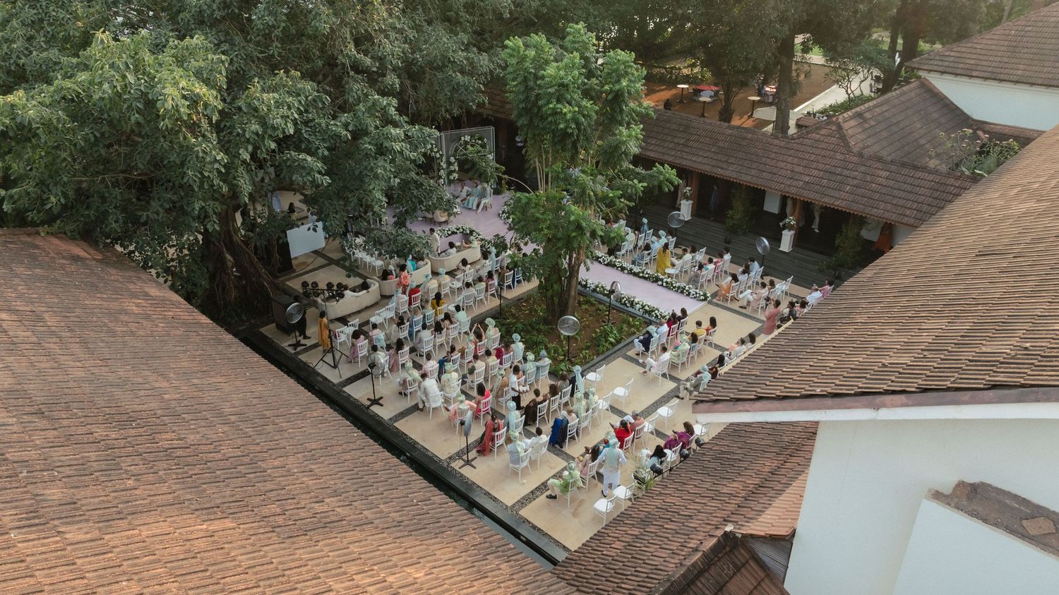 Aerial view of a large crowd gathered in a courtyard surrounded by lush trees and traditional tiled-roof buildings.