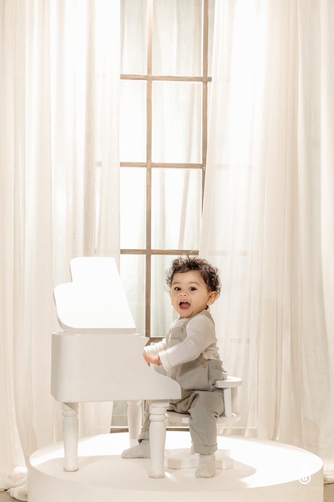 Cute toddler in gray outfit sitting at a small white grand piano by a bright window with sheer curtains.