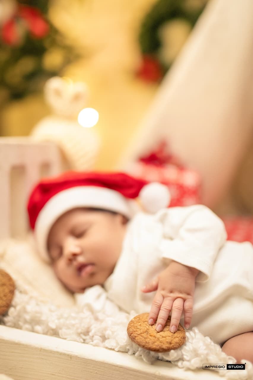 Sleeping newborn baby wearing a Santa hat reaches for a cookie, surrounded by Christmas decorations and warm bokeh lights.