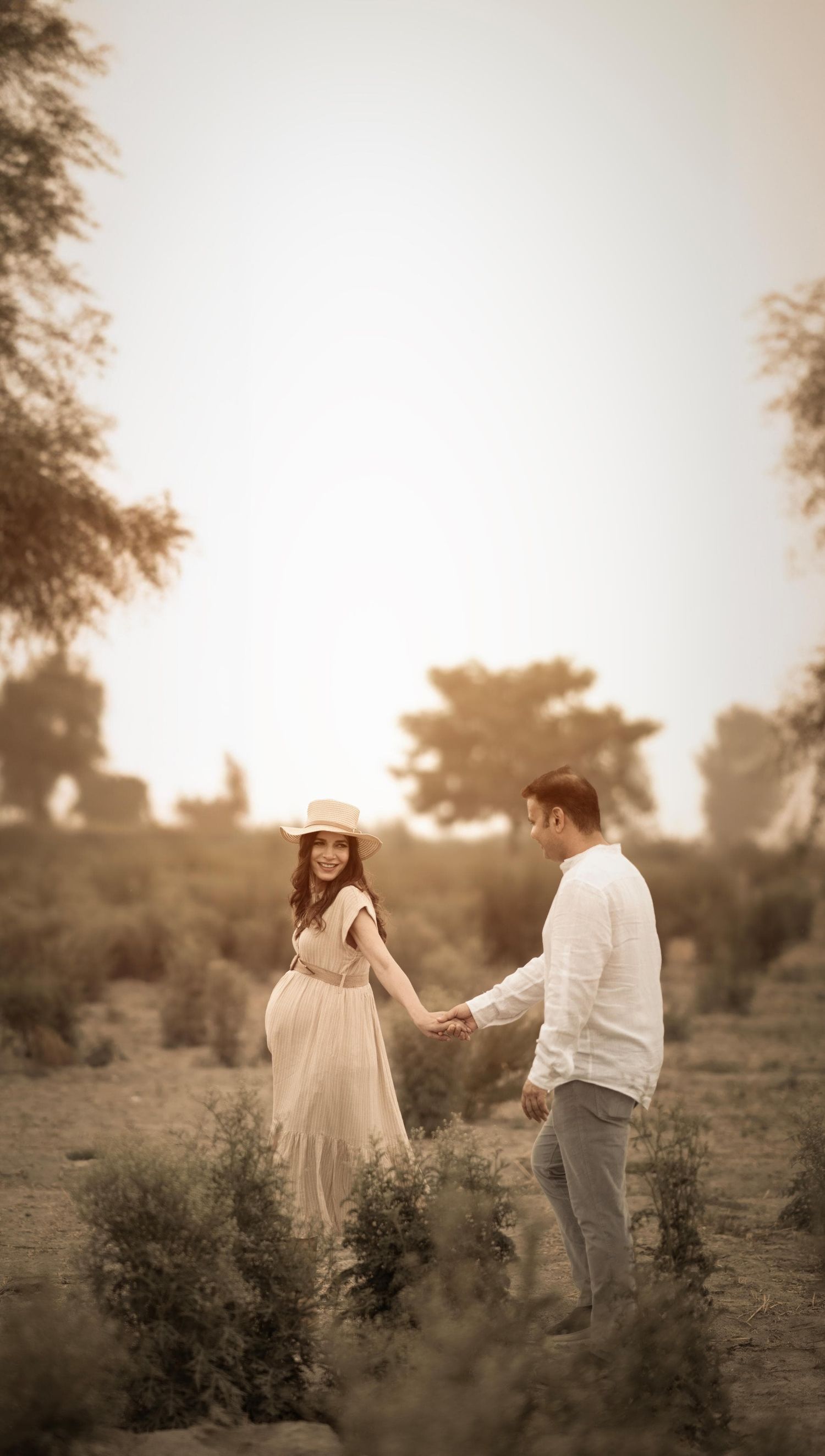 A couple holds hands in a golden-hued outdoor setting, woman in flowing dress and hat smiling at man in white shirt.