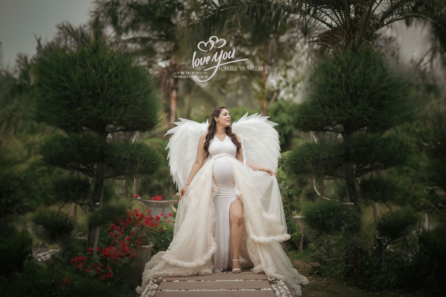 An ethereal maternity photo featuring white feathered wings and flowing gown in a garden setting.
