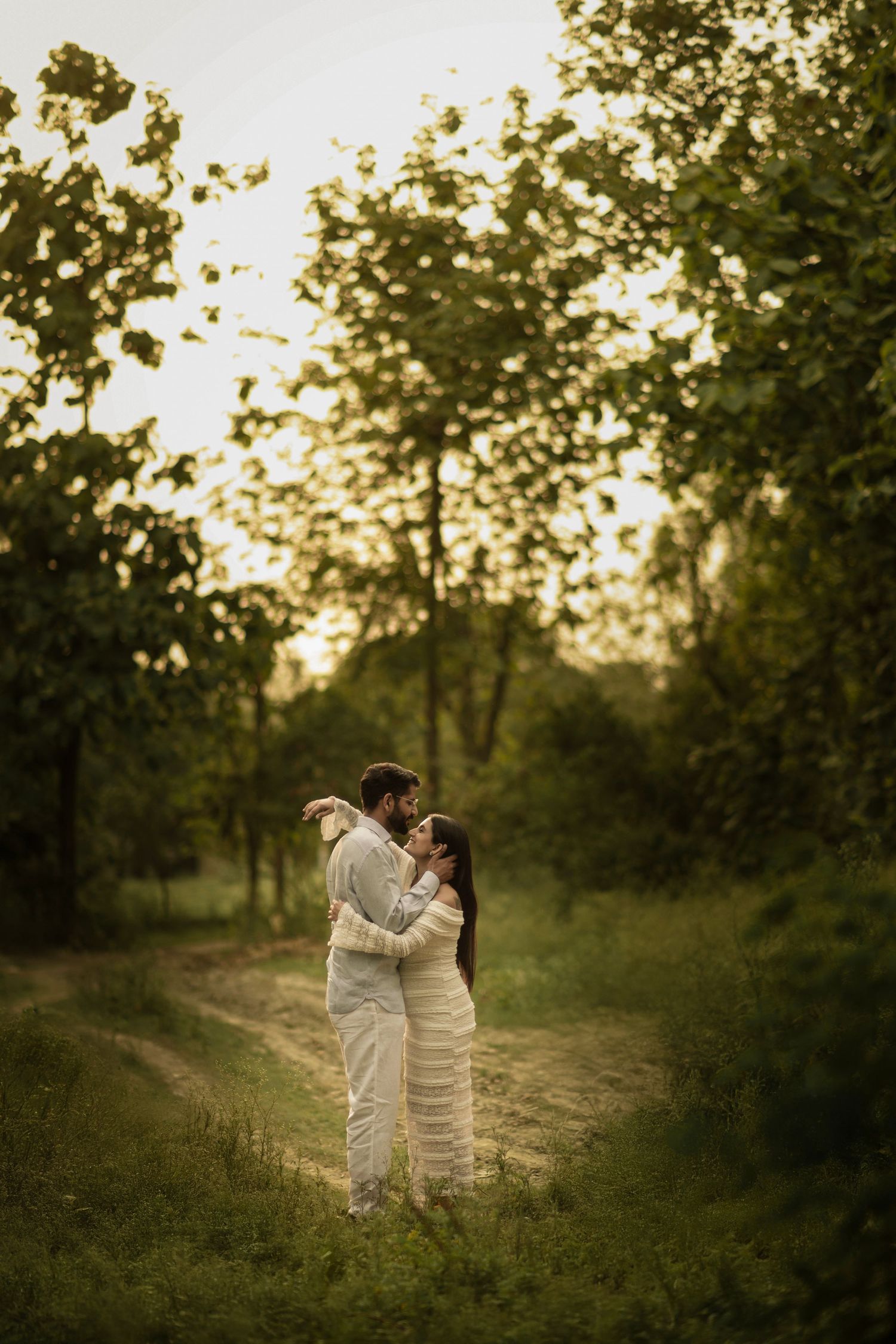 Two people in white outfits share an intimate moment on a sunlit path surrounded by trees at dusk.