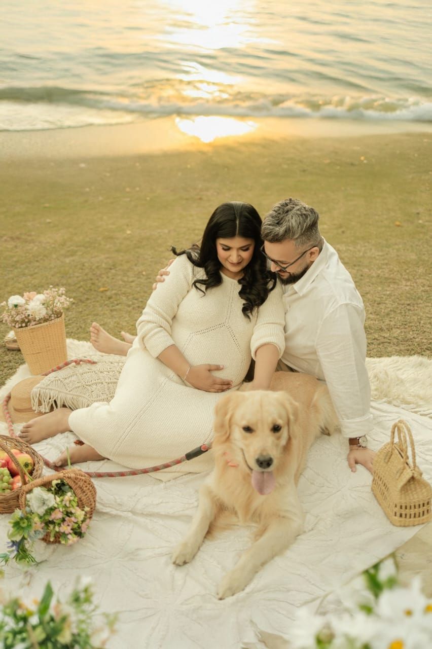 Pregnant couple sits on beach blanket at sunset with golden retriever dog, surrounded by flowers and picnic basket.
