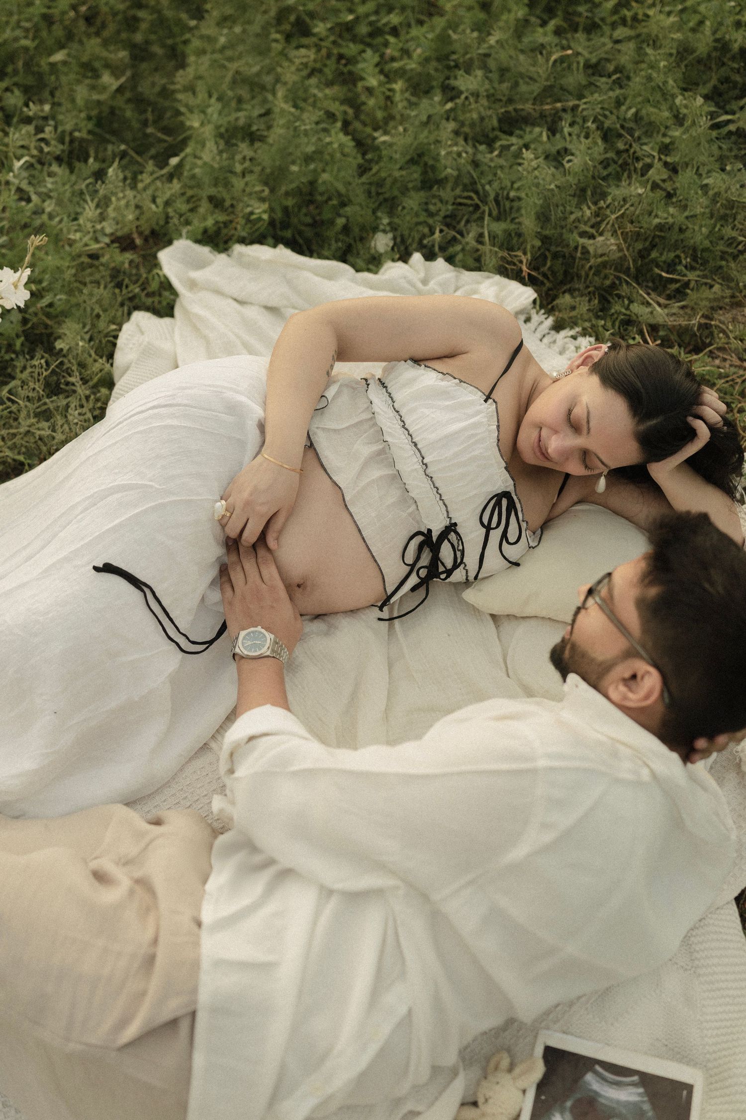 Pregnant woman in white dress lying on blanket in grass while partner rests head on her belly during maternity photoshoot.