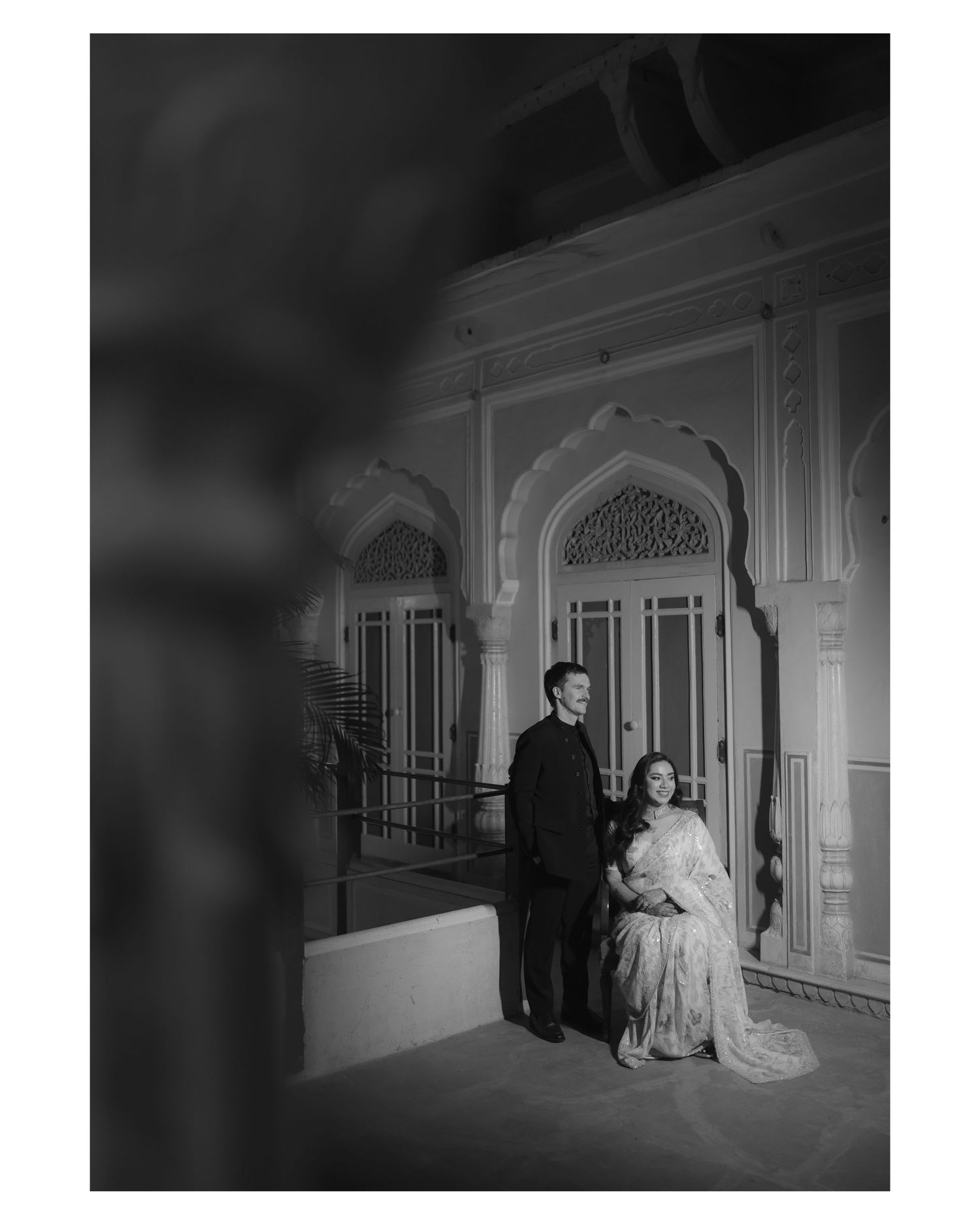 A black and white vintage photograph shows a couple in formal attire standing in an elegant hallway with ornate details.