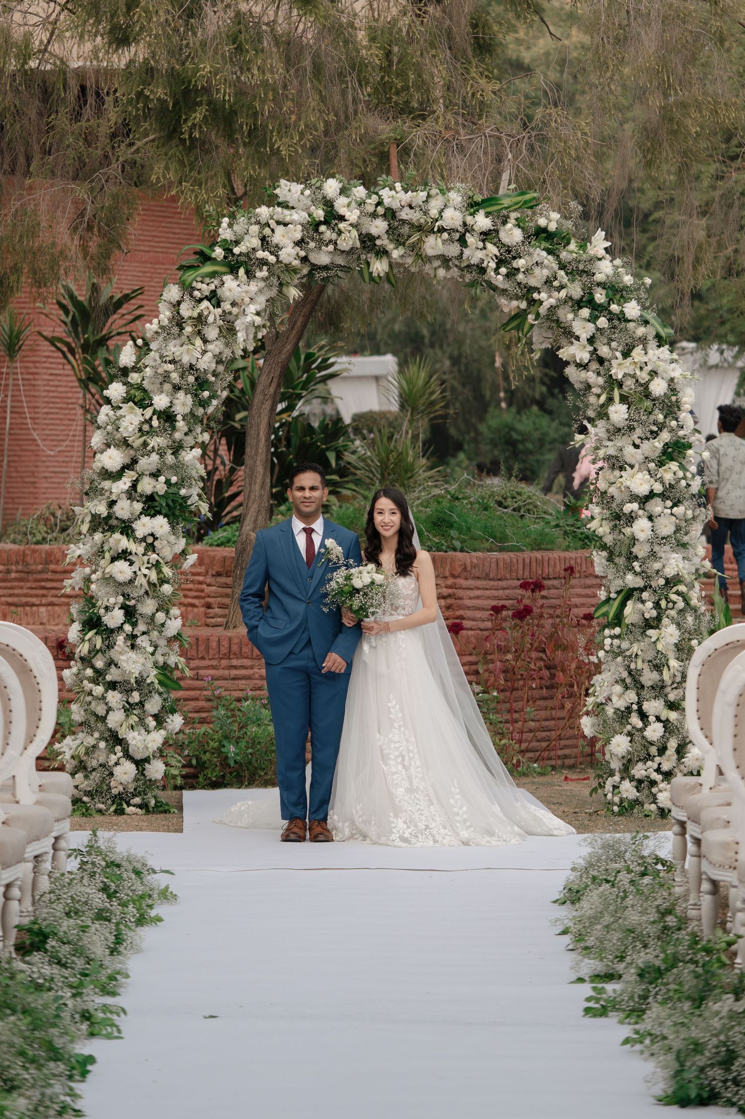 Elegant outdoor wedding ceremony setup with white floral arch and brick wall backdrop in a garden setting.