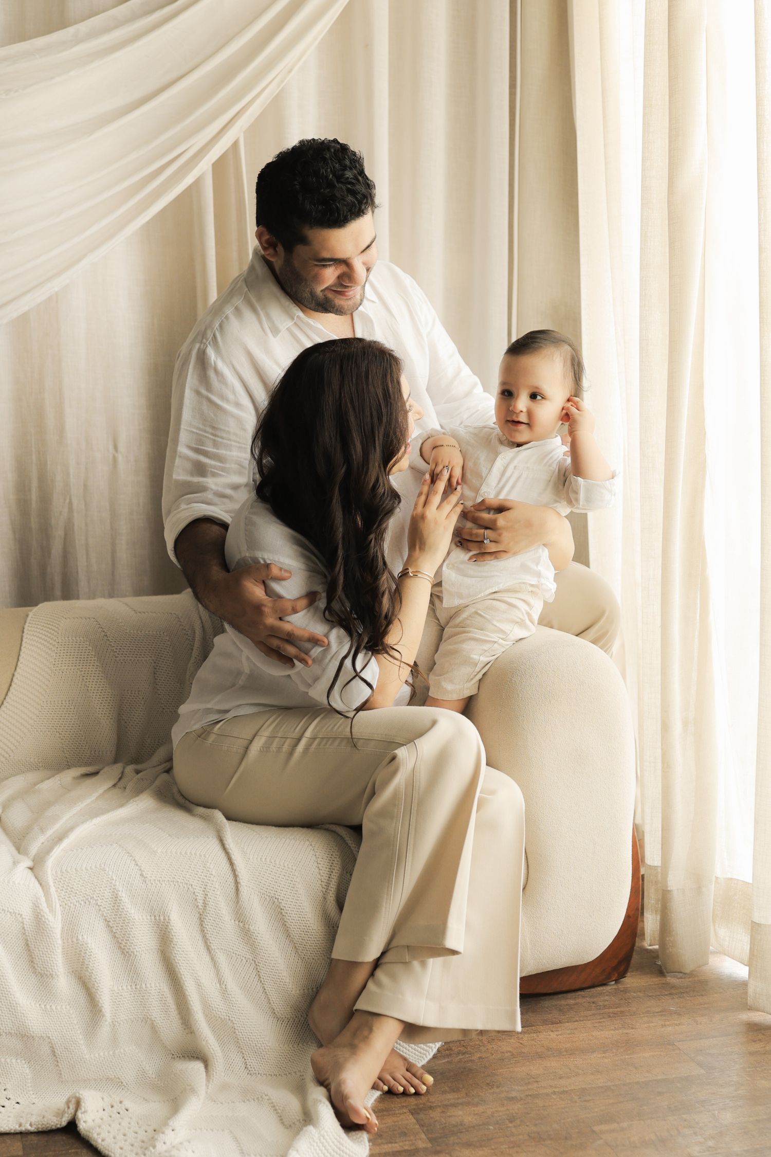 A happy family in white outfits sits on a beige sofa, parents smiling at their baby near sheer curtains.