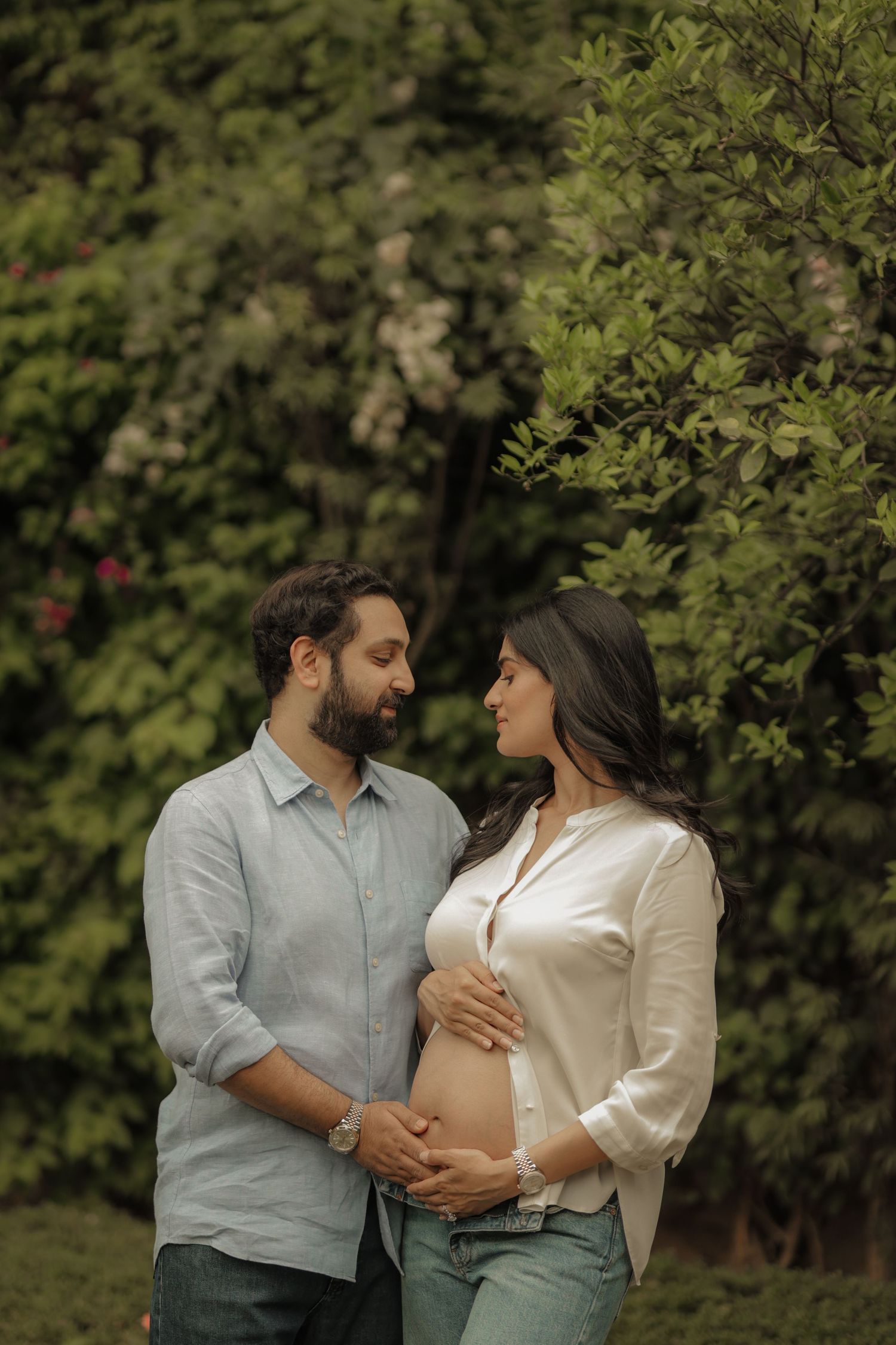 Couple sharing a tender moment outdoors, woman with baby bump in white shirt, man in blue, surrounded by lush green trees.