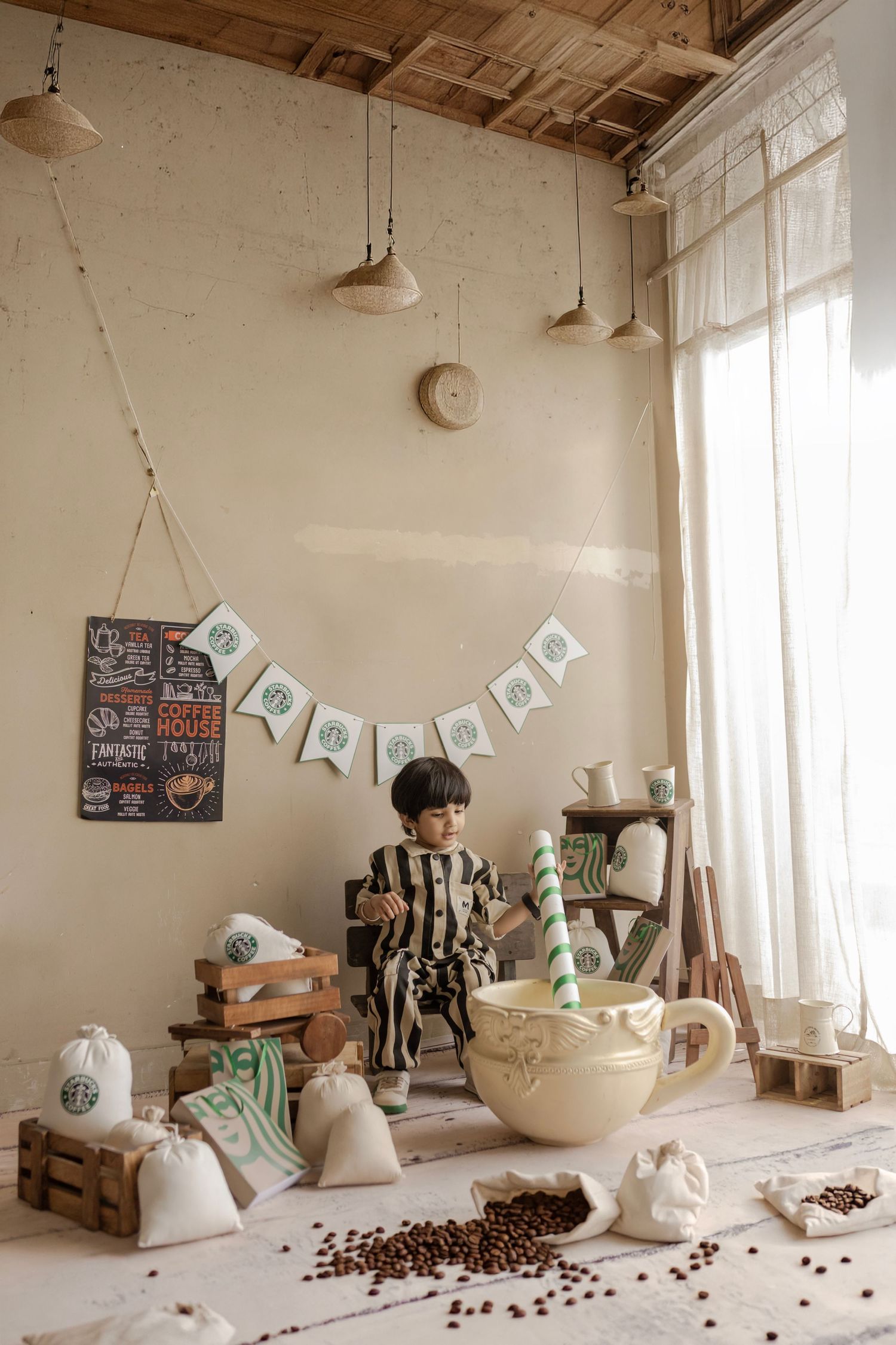 A child sits beside an oversized coffee cup prop surrounded by burlap sacks and coffee beans in a cozy cafe-themed photo setup.