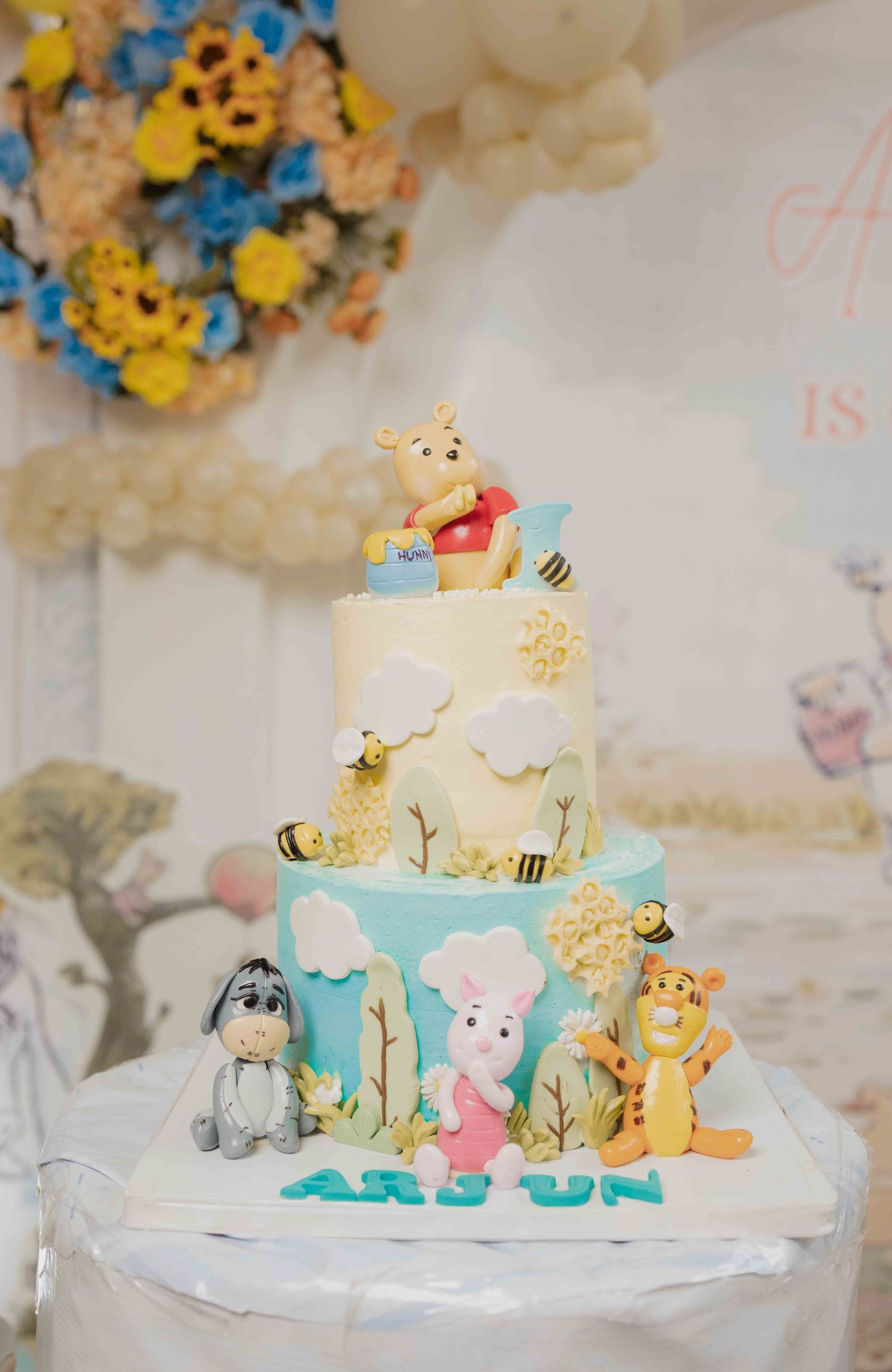 Two-tier Winnie the Pooh birthday cake with Eeyore, Piglet, and Tigger figurines, decorated for Arjun's celebration.