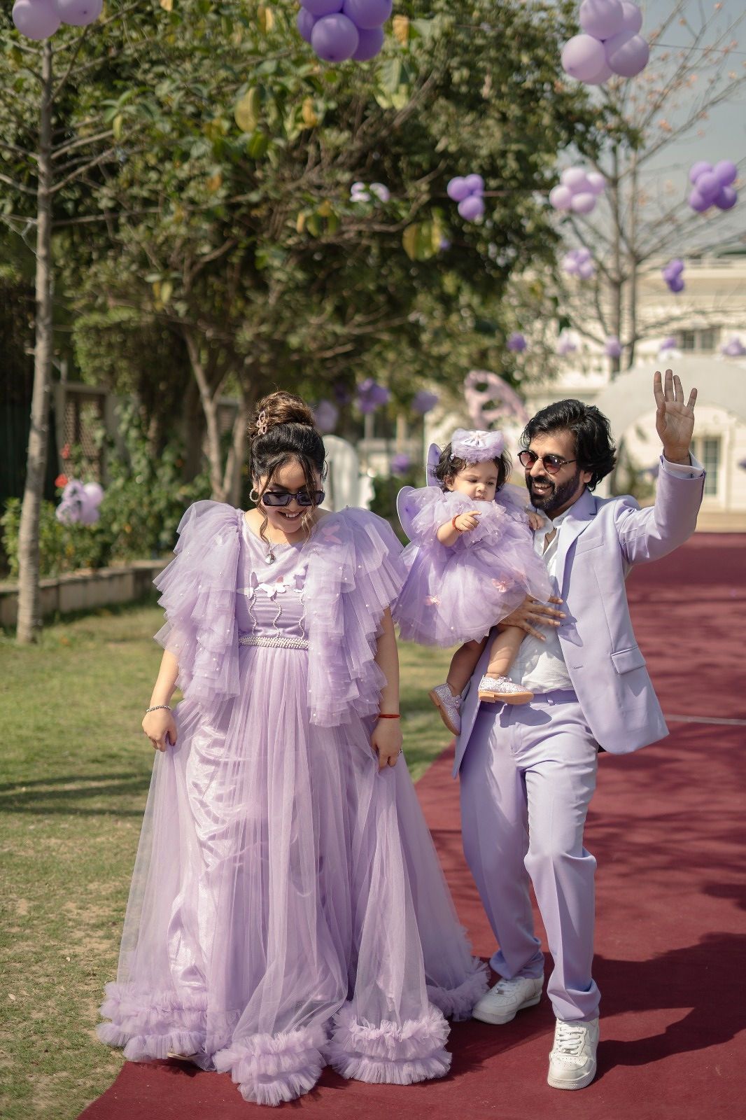 Family in matching lavender outfits at an outdoor celebration with purple balloons floating above a red carpet.