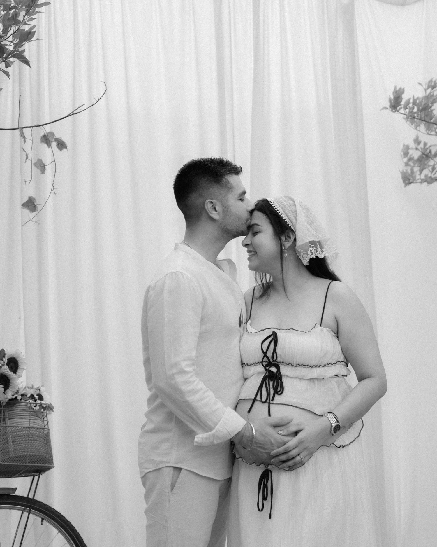 Black and white maternity photo of a couple in white outfits, man kissing pregnant womans forehead.
