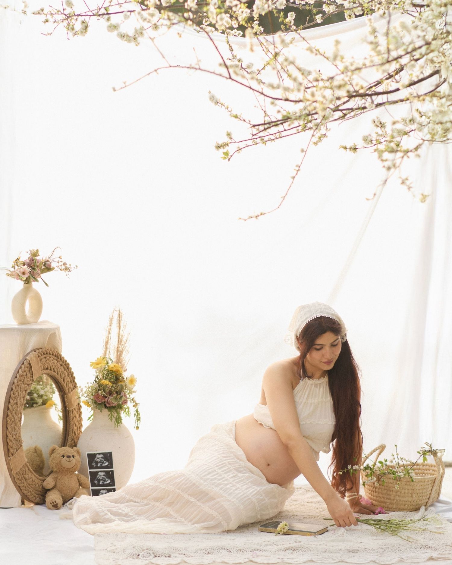 Pregnant woman in white lace dress sits gracefully under cherry blossoms in a serene, floral-decorated maternity photo shoot.