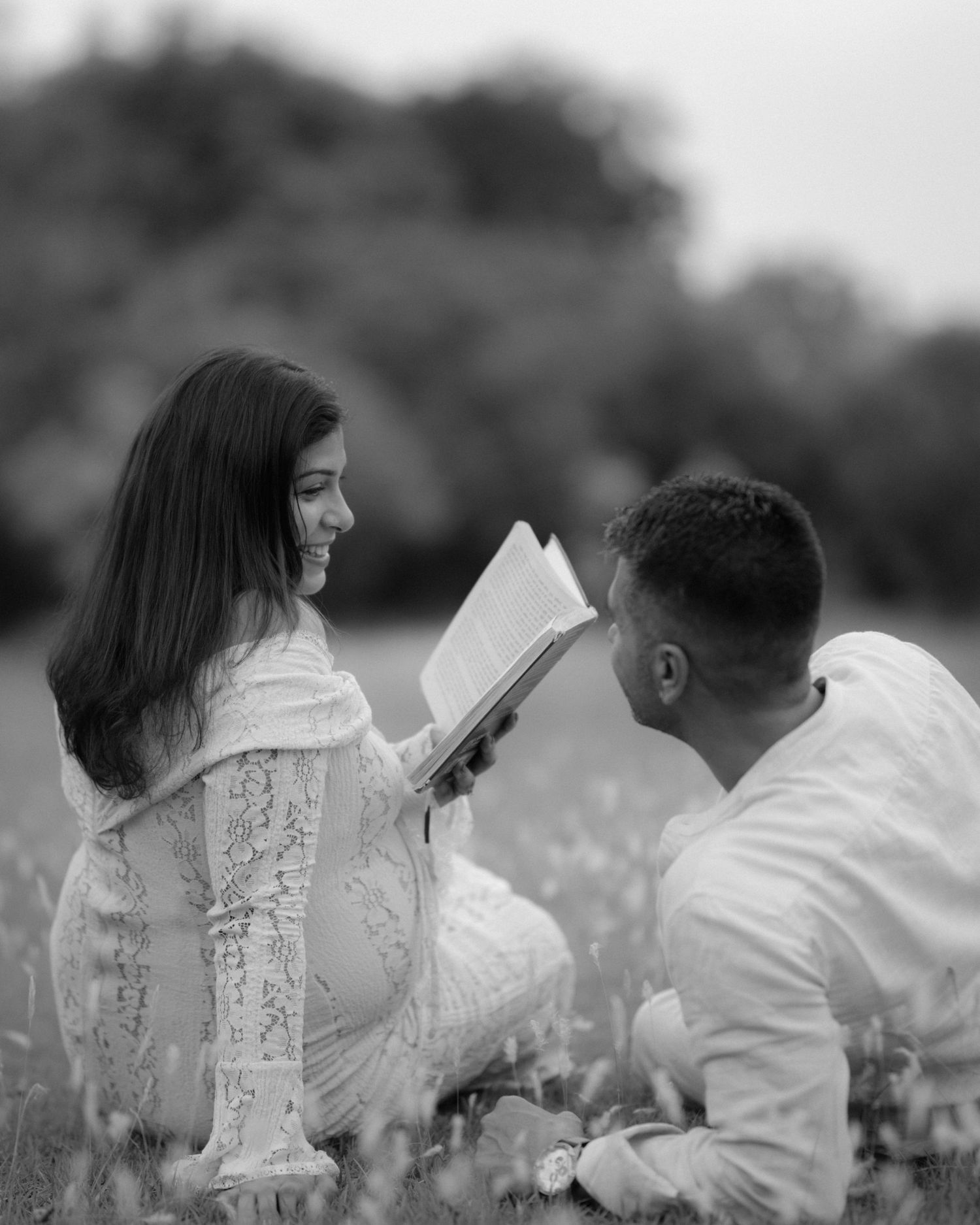Black and white photo of a couple sitting in a field, woman in lace dress smiling while reading a book to her partner.