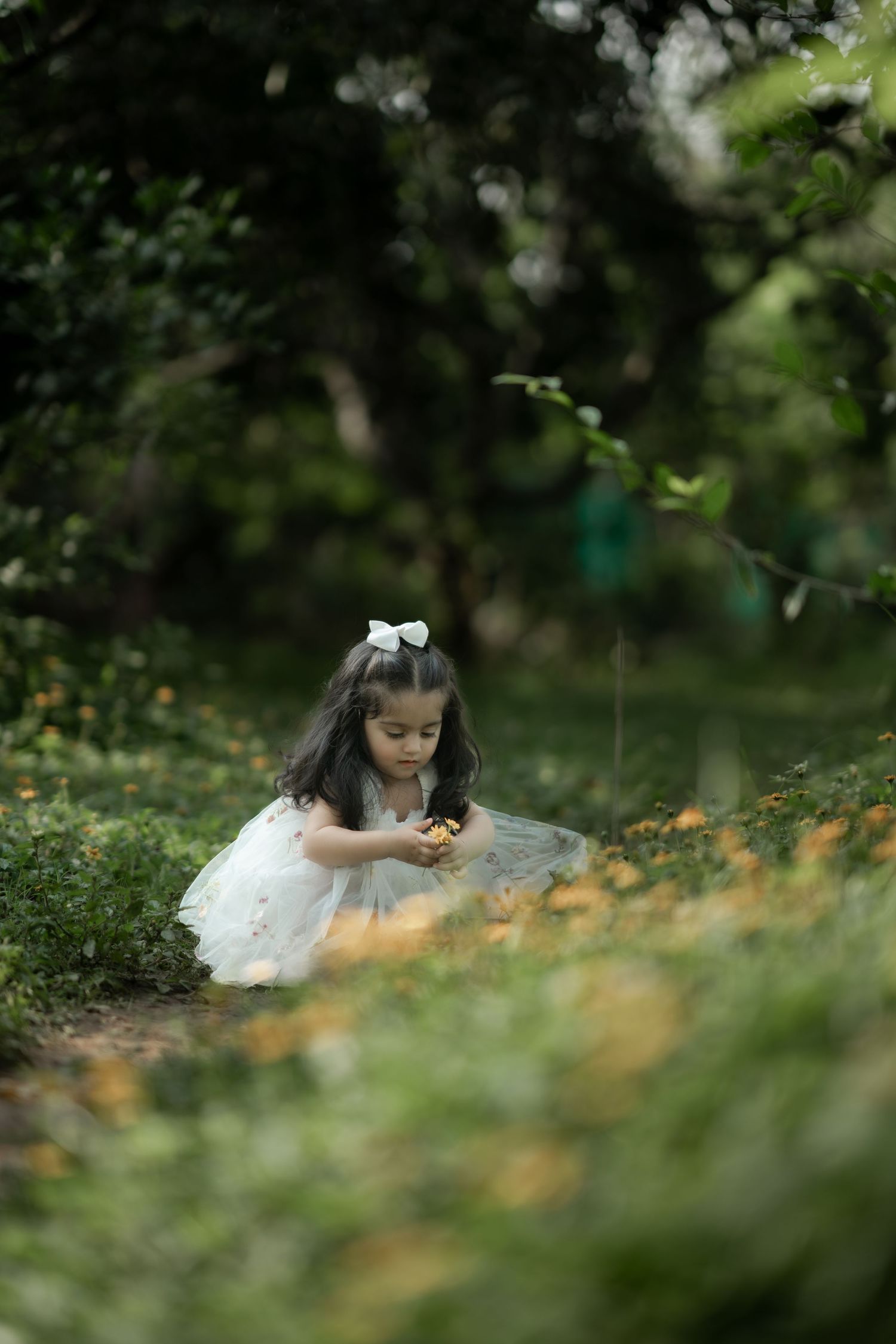 A young girl in a white tulle dress and bow picks yellow flowers in a lush green garden.