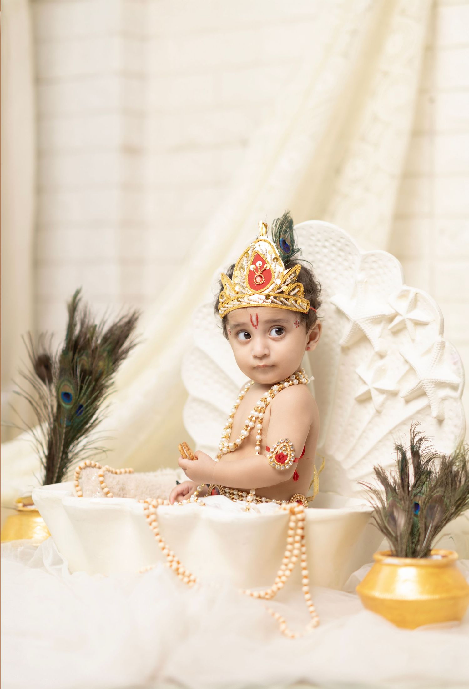 Baby dressed as Lord Krishna with crown, pearls, and peacock feathers in a decorative white shell setup.
