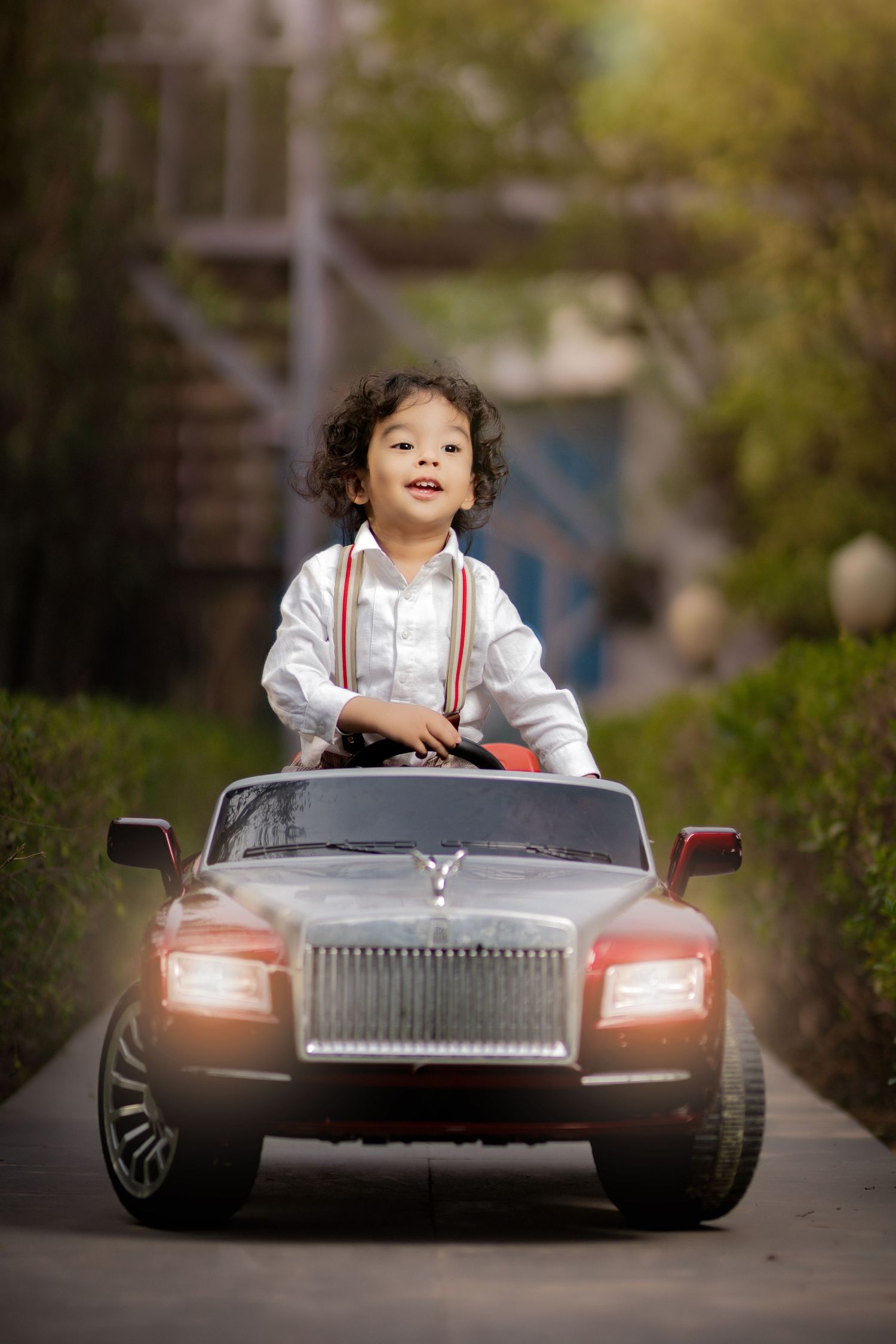 Young boy in white shirt with suspenders smiling while riding a toy Rolls-Royce electric car outdoors.