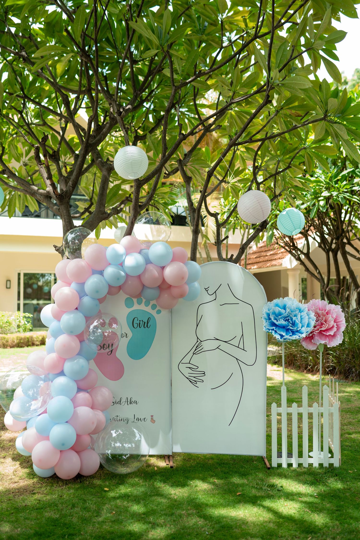 Outdoor baby shower decor with pink and blue balloon arch, pregnant woman illustration, and paper flowers in garden.