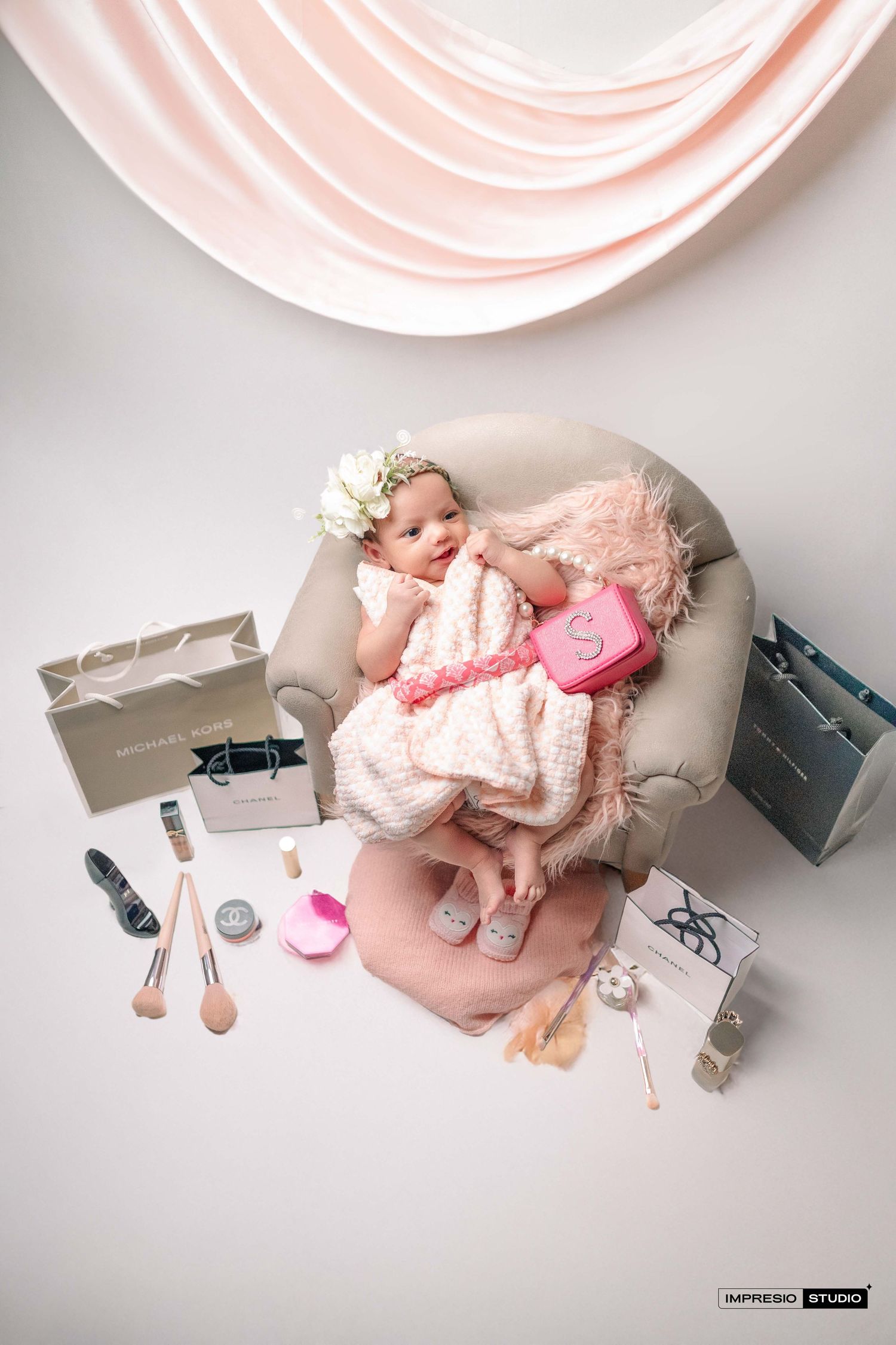 A baby girl in a pink dress sits in a mini chair surrounded by makeup, shopping bags, and accessories in a studio photo.