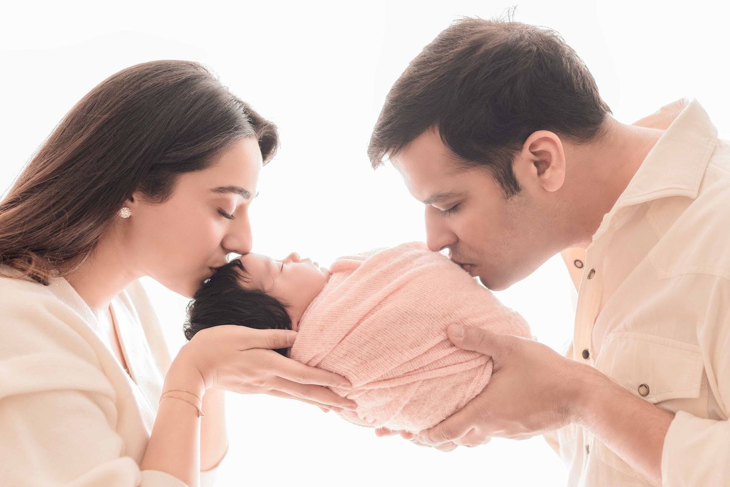 Parents tenderly kissing their newborn baby in a soft, bright studio portrait.