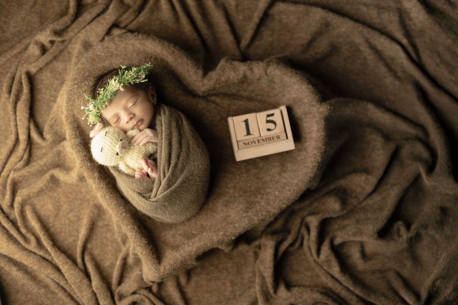 Newborn baby wrapped in brown swaddle with floral crown, sleeping in heart-shaped blanket with November 15 date blocks.