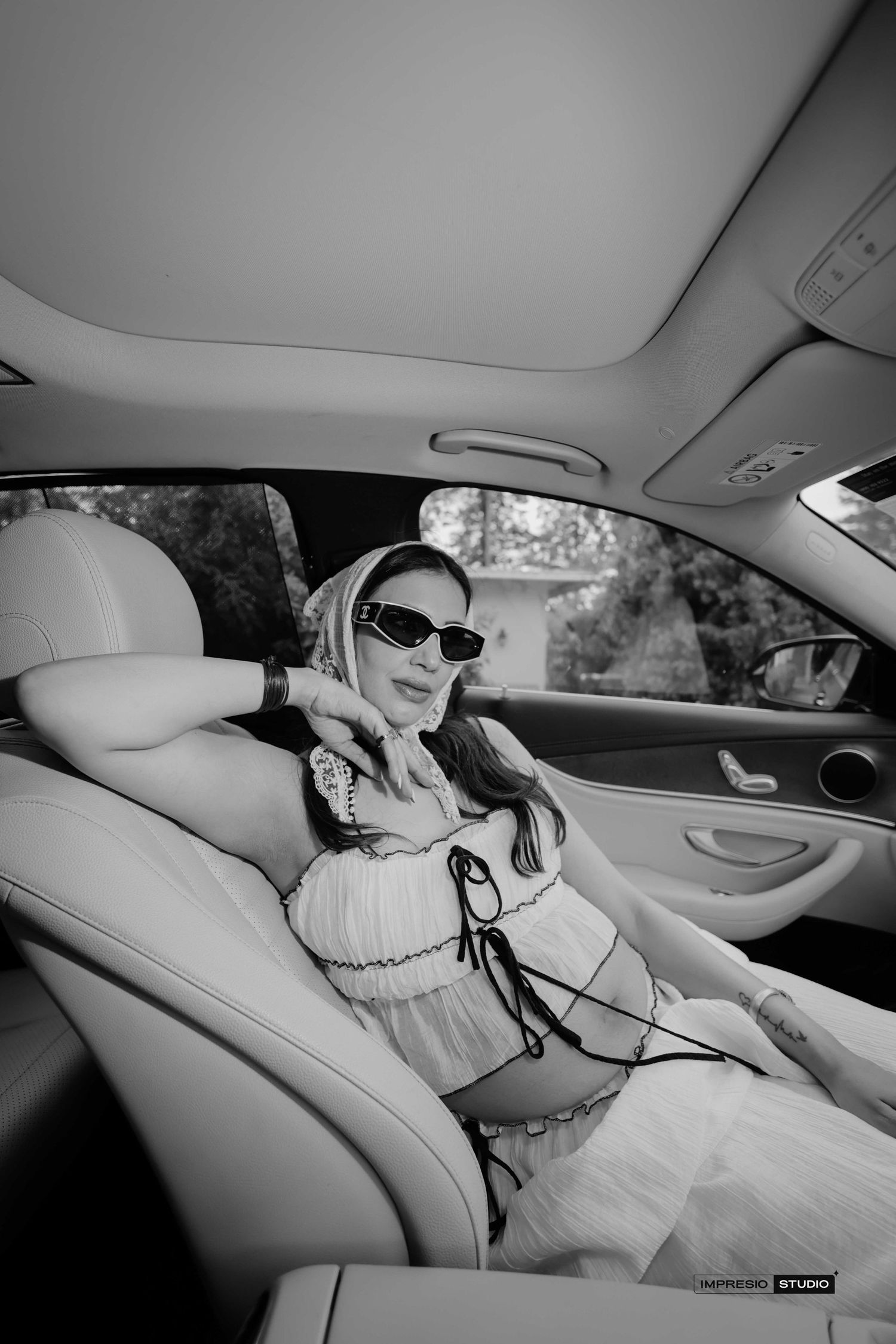 A stylish woman in sunglasses and a white outfit relaxes in the backseat of a luxury car in black and white.