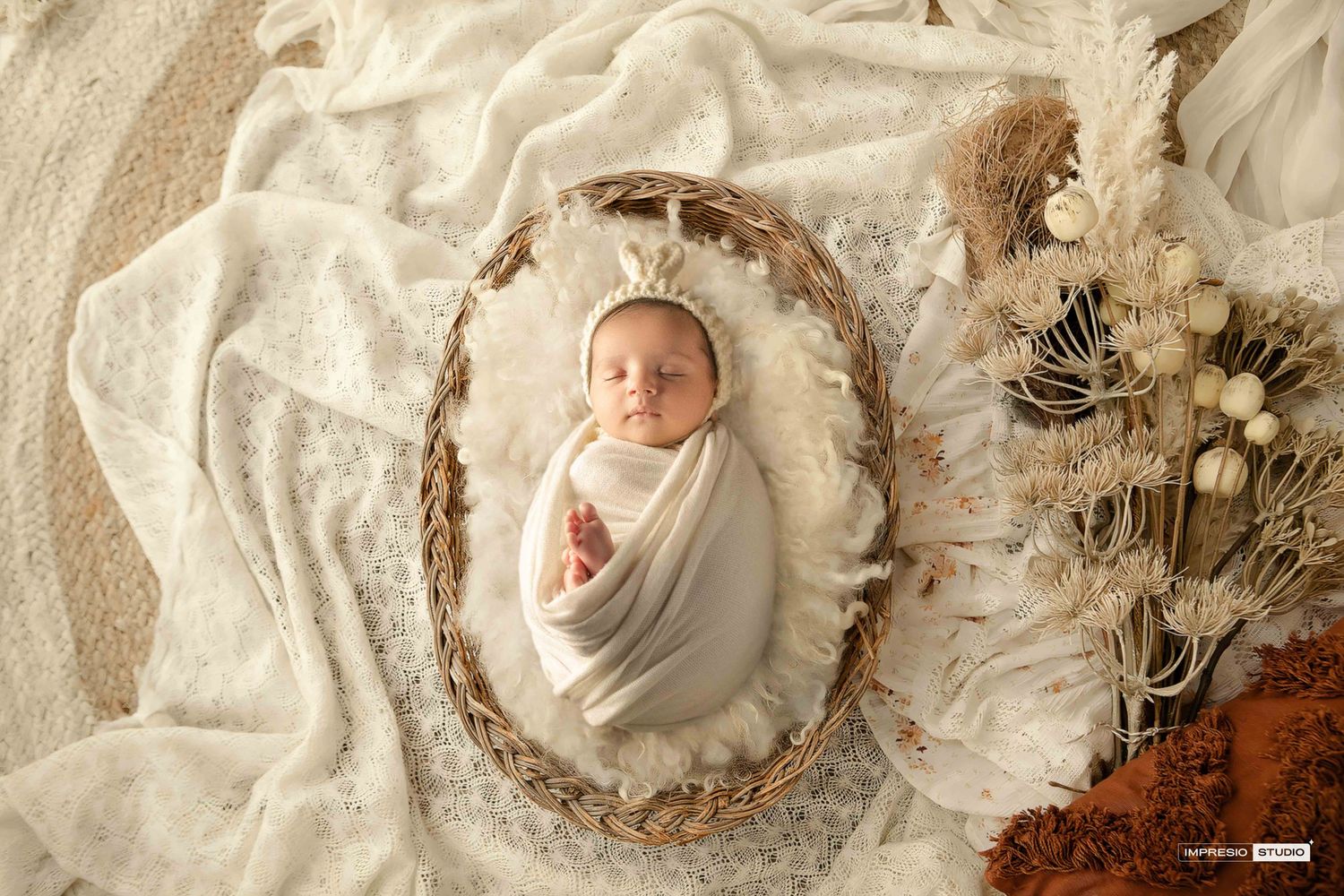 Newborn baby swaddled in white wrap, nestled in wicker bowl with dried flowers on cream lace backdrop.