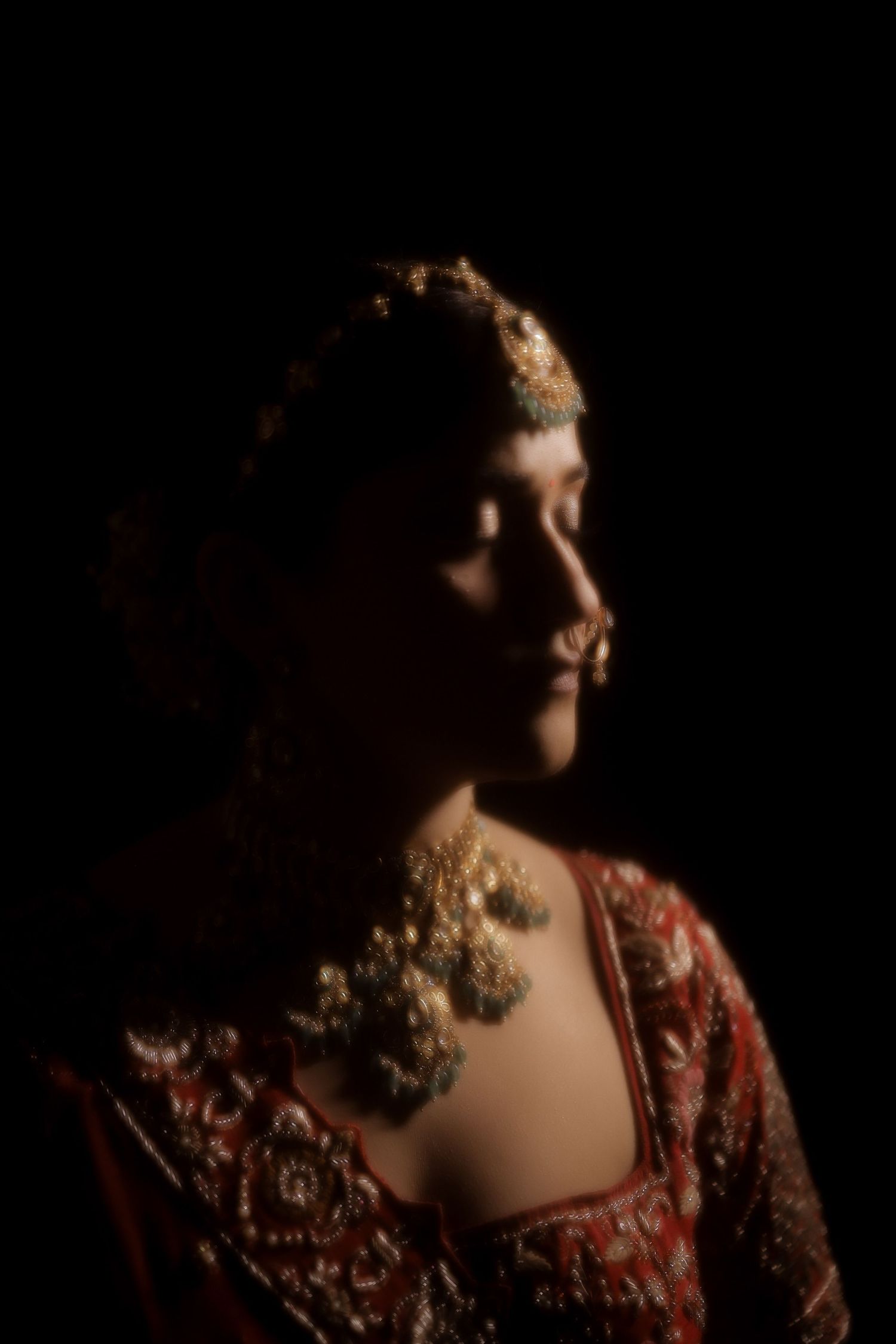 A woman in traditional South Asian bridal attire with gold jewelry and red embroidered dress poses in dramatic low-key lighting.