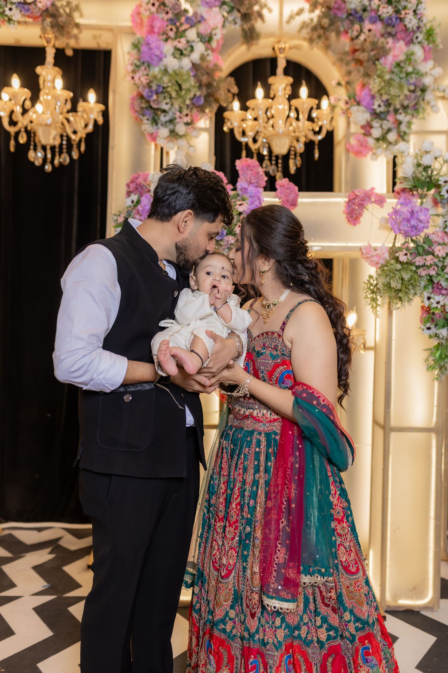 Parents in a black vest and a teal and red embroidered lehenga gently kiss their baby under floral arrangements and crystal chandeliers.
