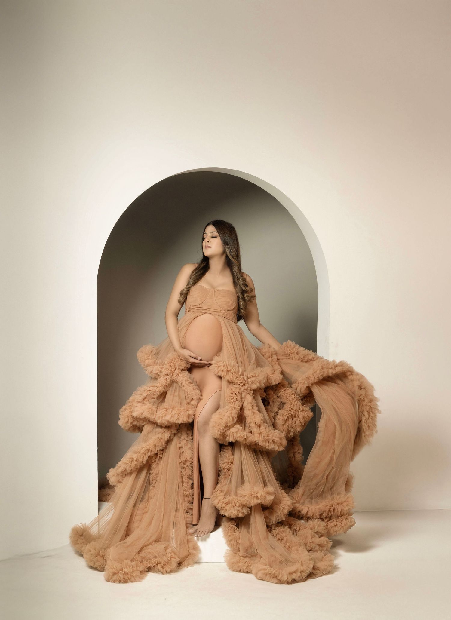 Dramatic maternity portrait with cascading ruffled dusty rose gown in studio arch setting.