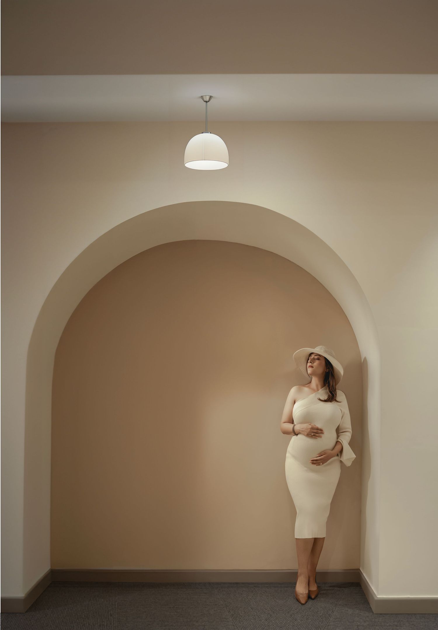 Pregnant woman in white dress and hat poses gracefully under arched alcove with pendant light.