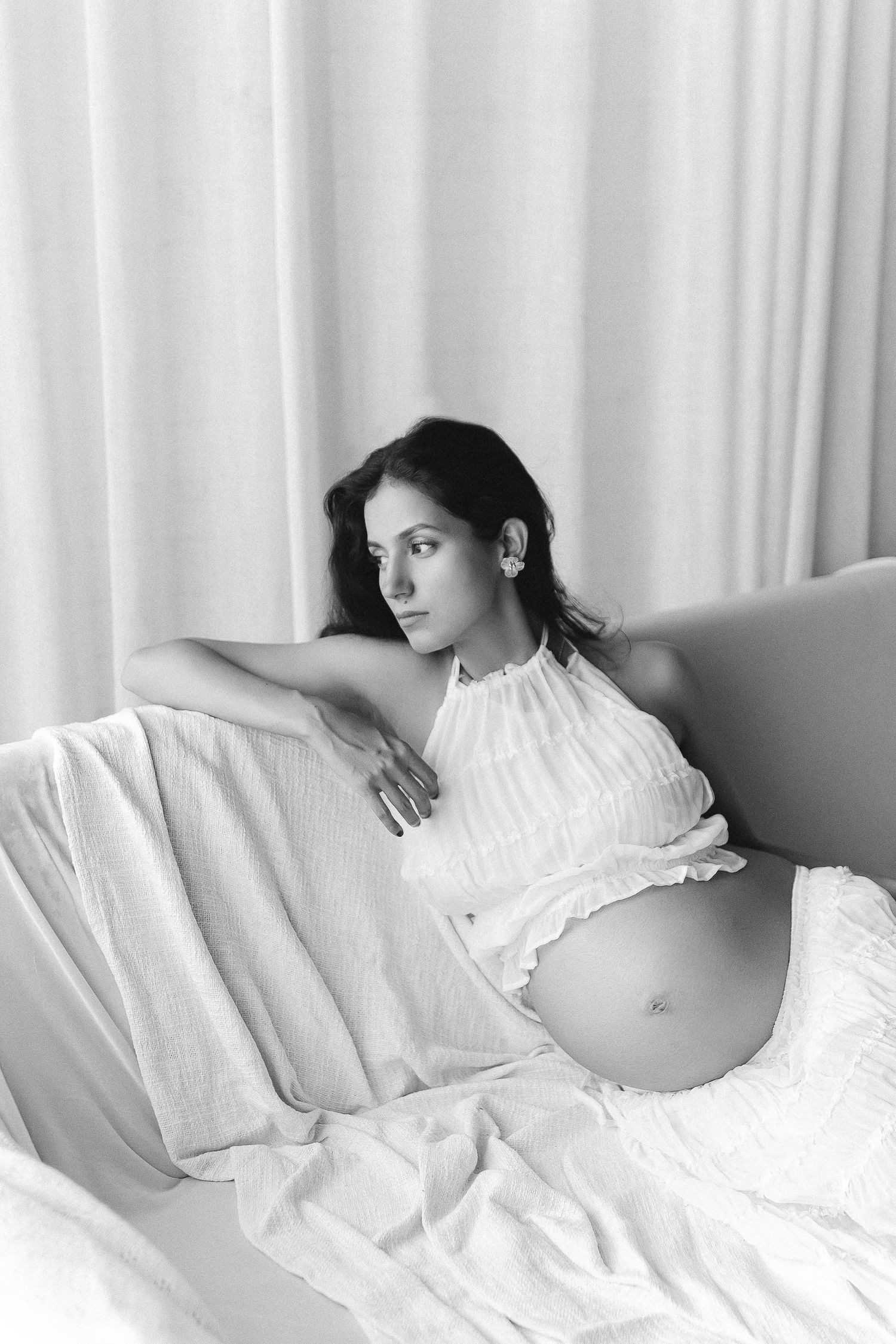 Black and white maternity photo of someone reclining on white couch wearing halter top showing pregnant belly.