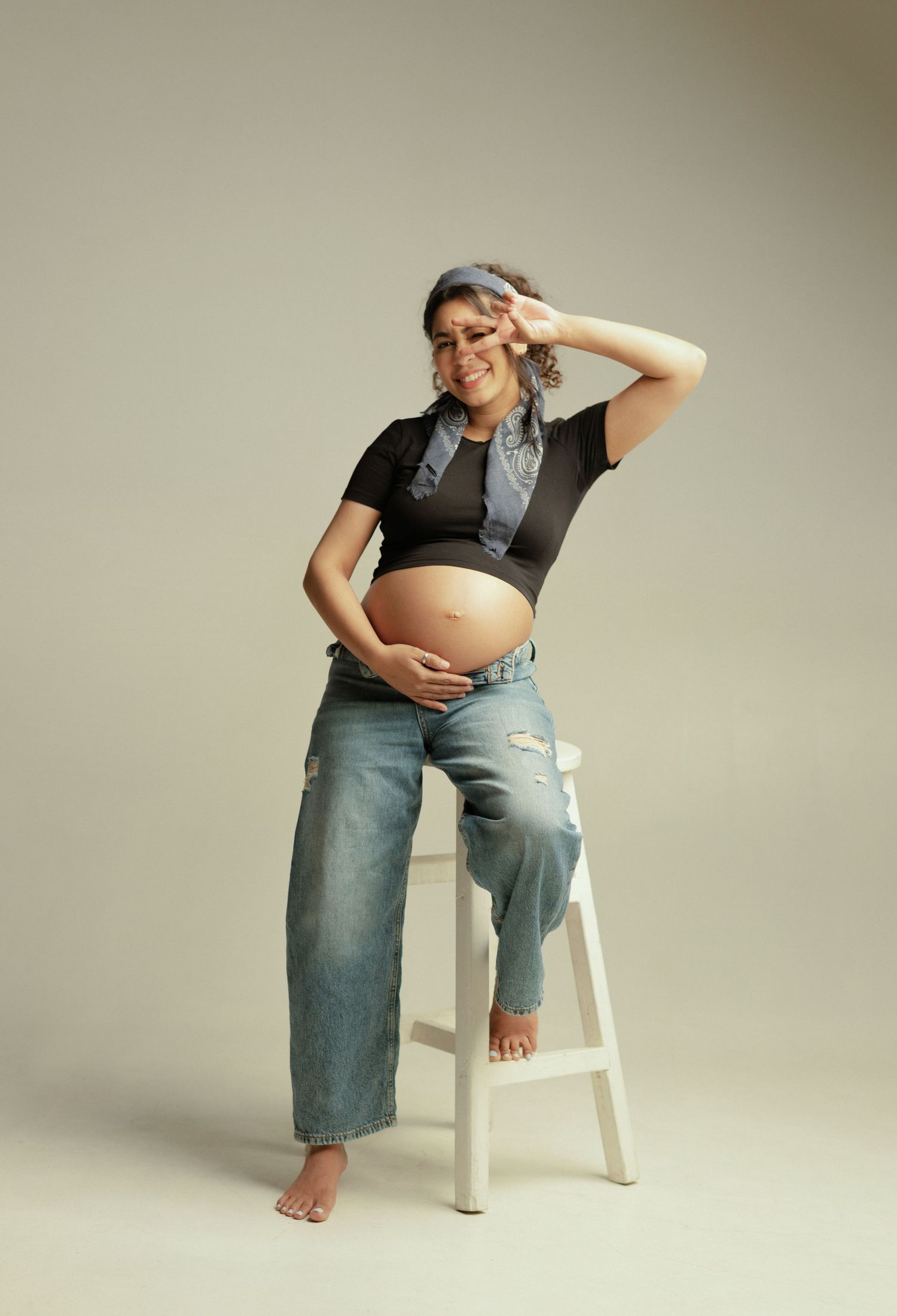 A person in denim overalls sits casually on a white chair against a neutral backdrop in a minimalist maternity photo series.