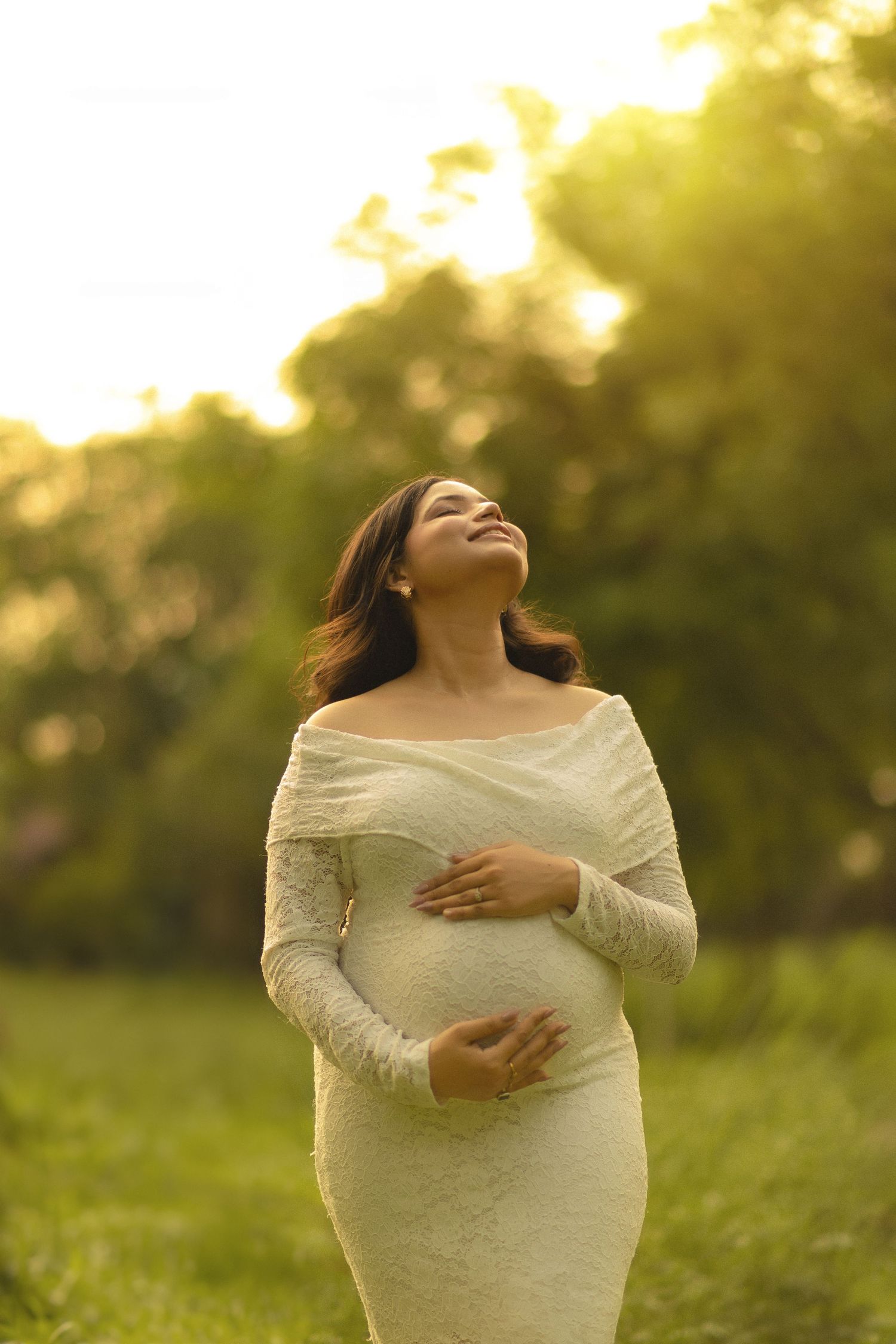Pregnant woman in white lace dress cradling her baby bump outdoors in golden sunlight, smiling joyfully.