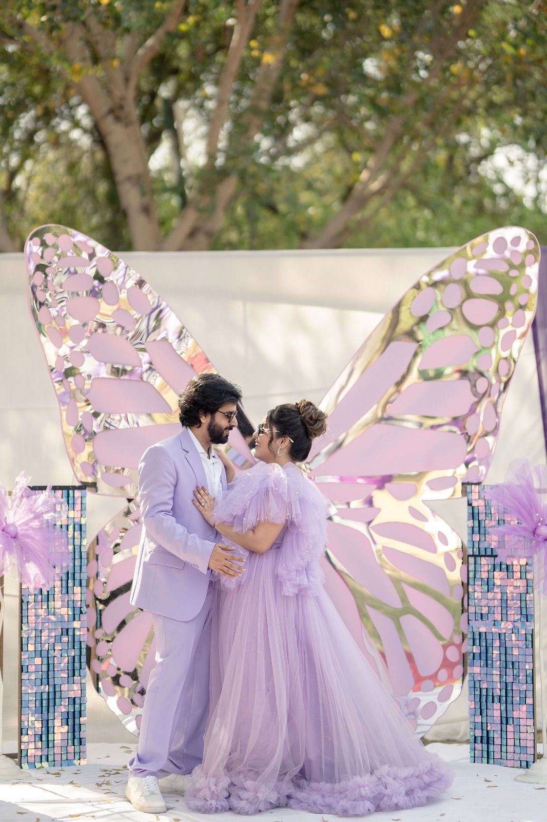 Couple in matching lavender outfits posing before a large decorative butterfly backdrop outdoors.
