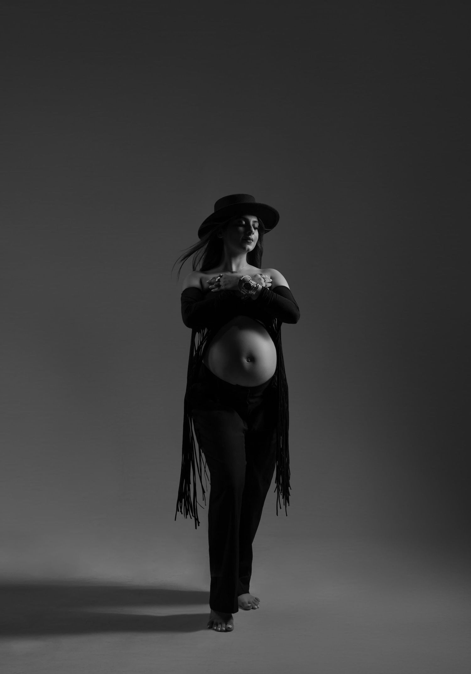 Black and white maternity photo of pregnant woman in hat and fringe jacket posing in studio.
