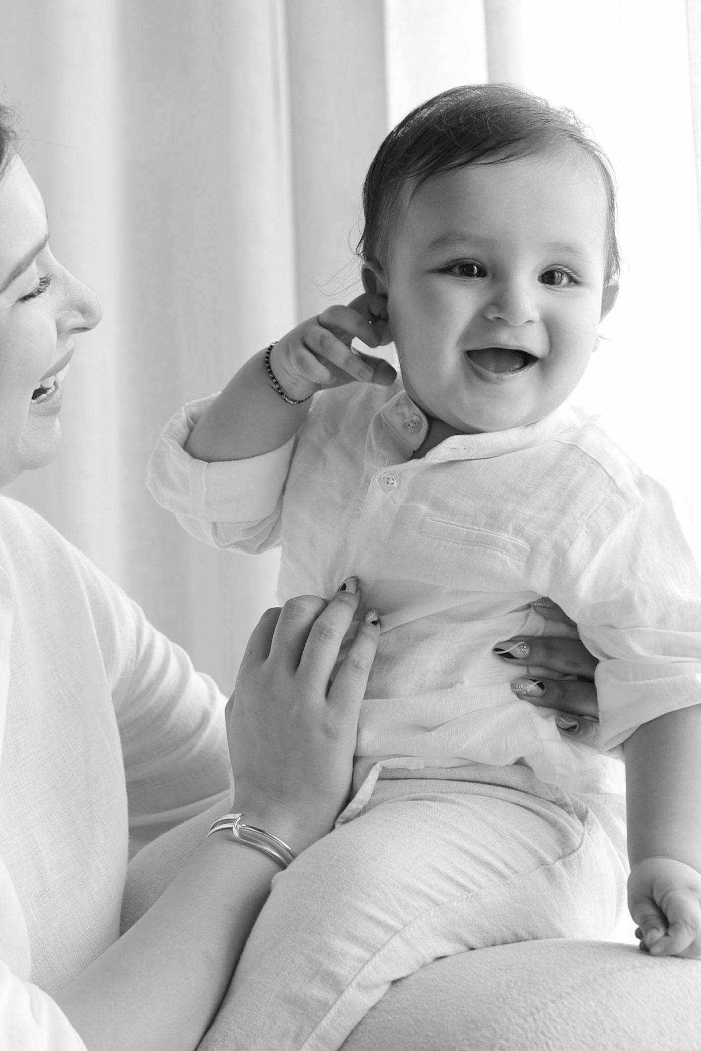 Black and white photo of a joyful baby in white outfit laughing while being held by a smiling adult.