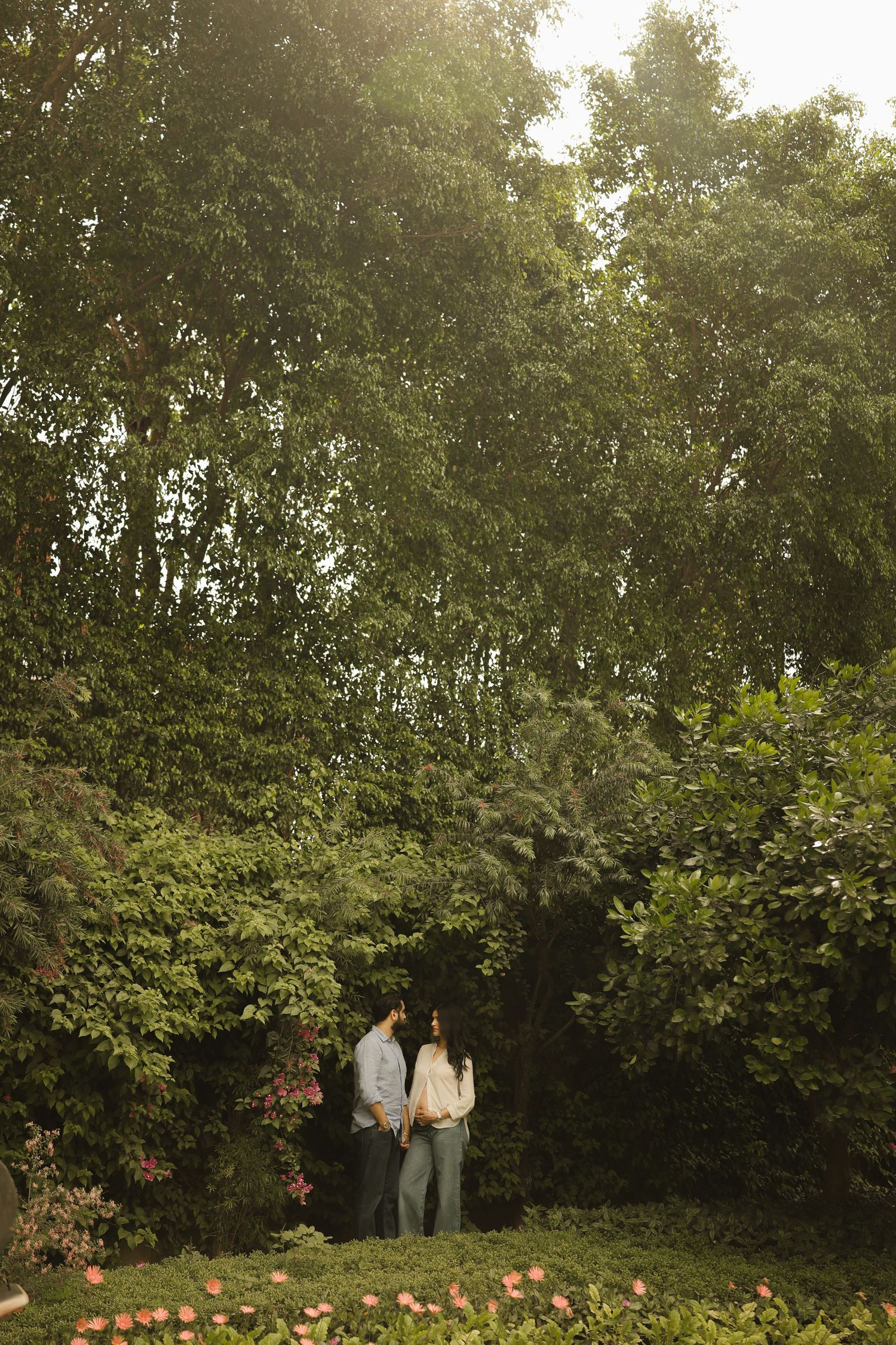 Couple standing hand in hand surrounded by lush green trees and pink flowers in a romantic garden setting.