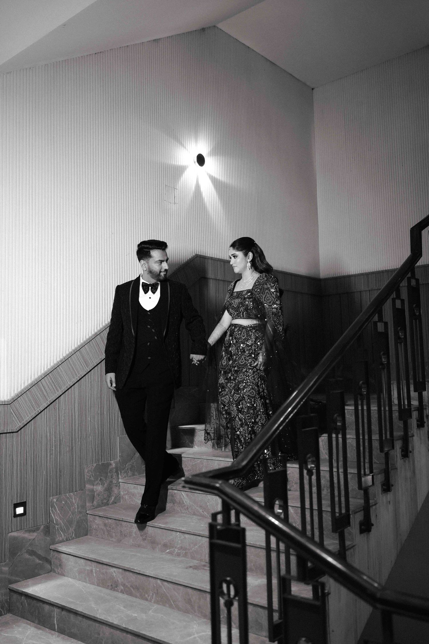 Elegant couple in formal attire holding hands on a grand staircase in a black and white wedding photo.