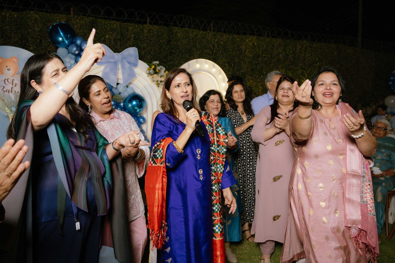 Women in colorful traditional South Asian attire singing and dancing together at an outdoor celebration event.