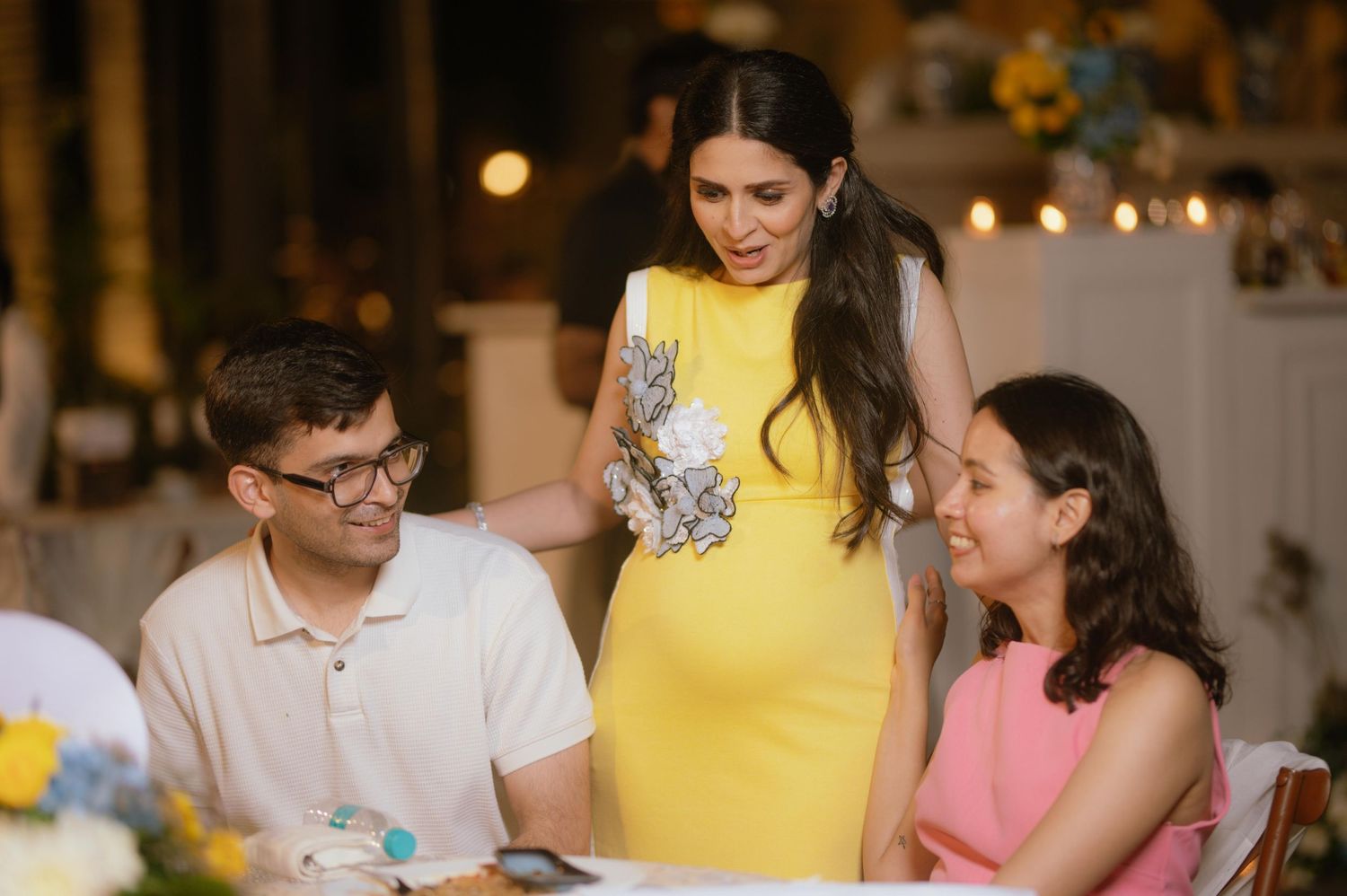 Woman in yellow dress smiles at two seated guests at an elegant evening event.