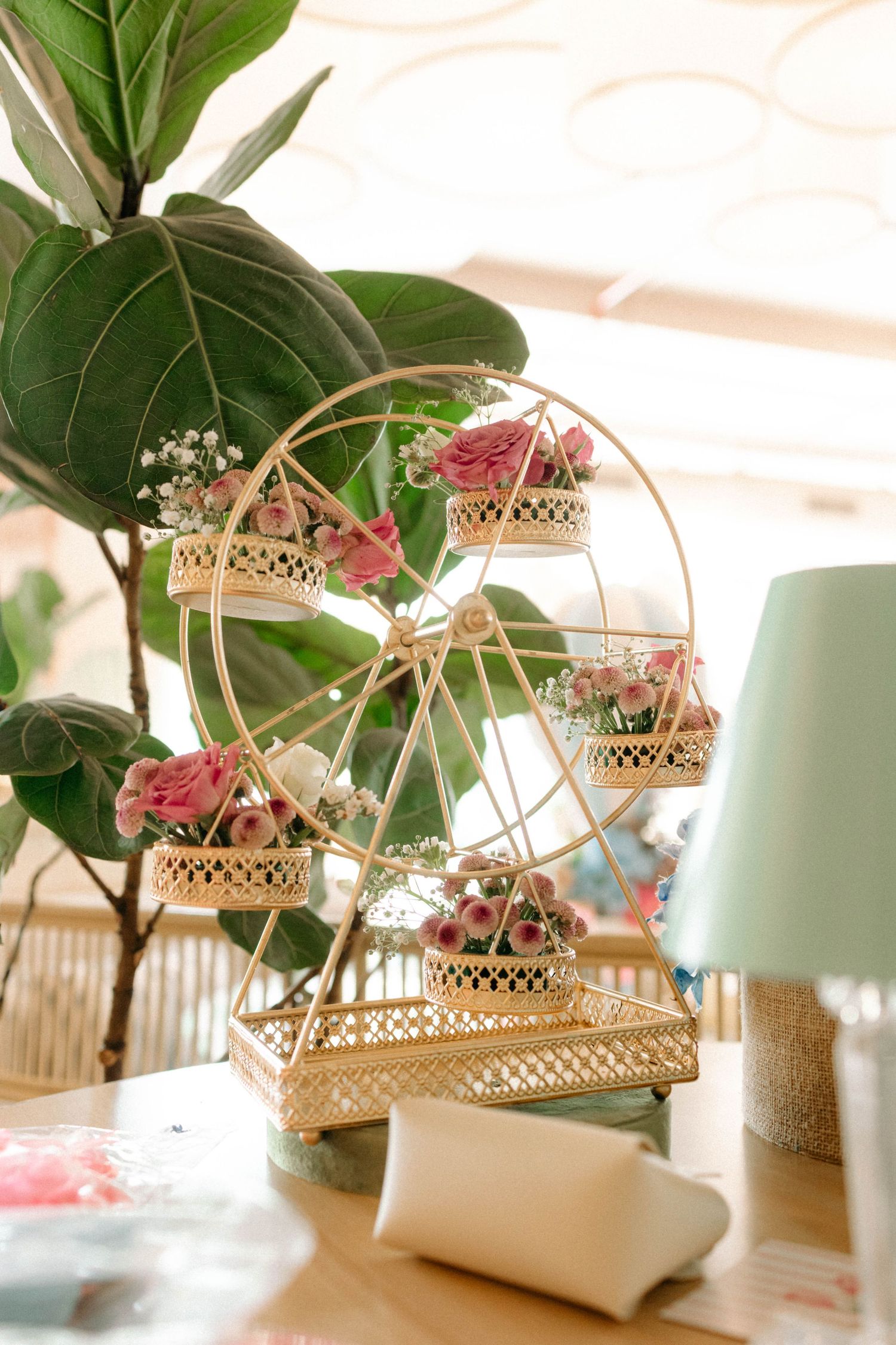 Gold ferris wheel centerpiece with pink roses in wicker baskets, displayed on a wooden table near a fiddle leaf fig plant.