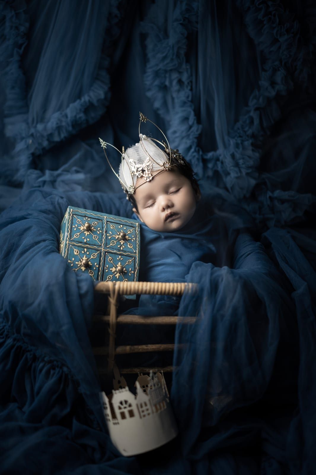 Sleeping newborn baby wearing a delicate crown, wrapped in blue tulle fabric with ornate decorative props in a royal-themed portrait.