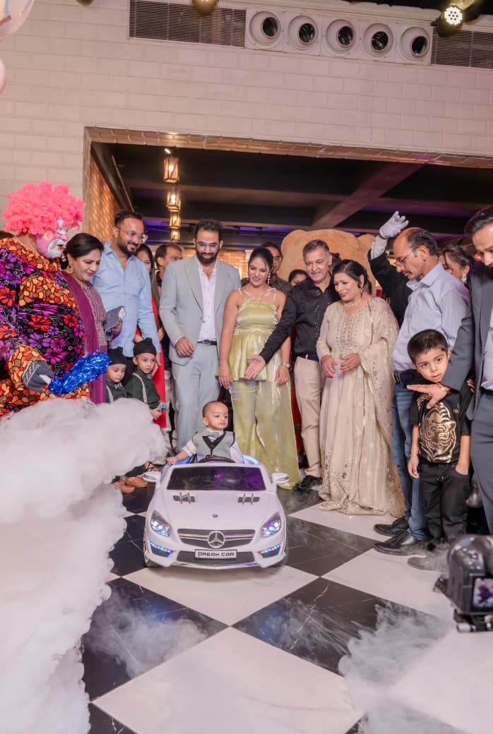 A baby sits in a toy Mercedes car surrounded by family and a clown at a festive first birthday party celebration.