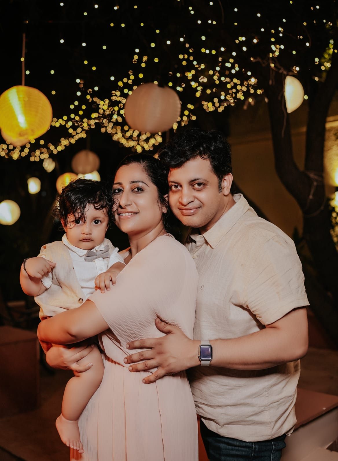A smiling couple holding a baby dressed in white at a festive outdoor party with glowing lanterns and string lights.