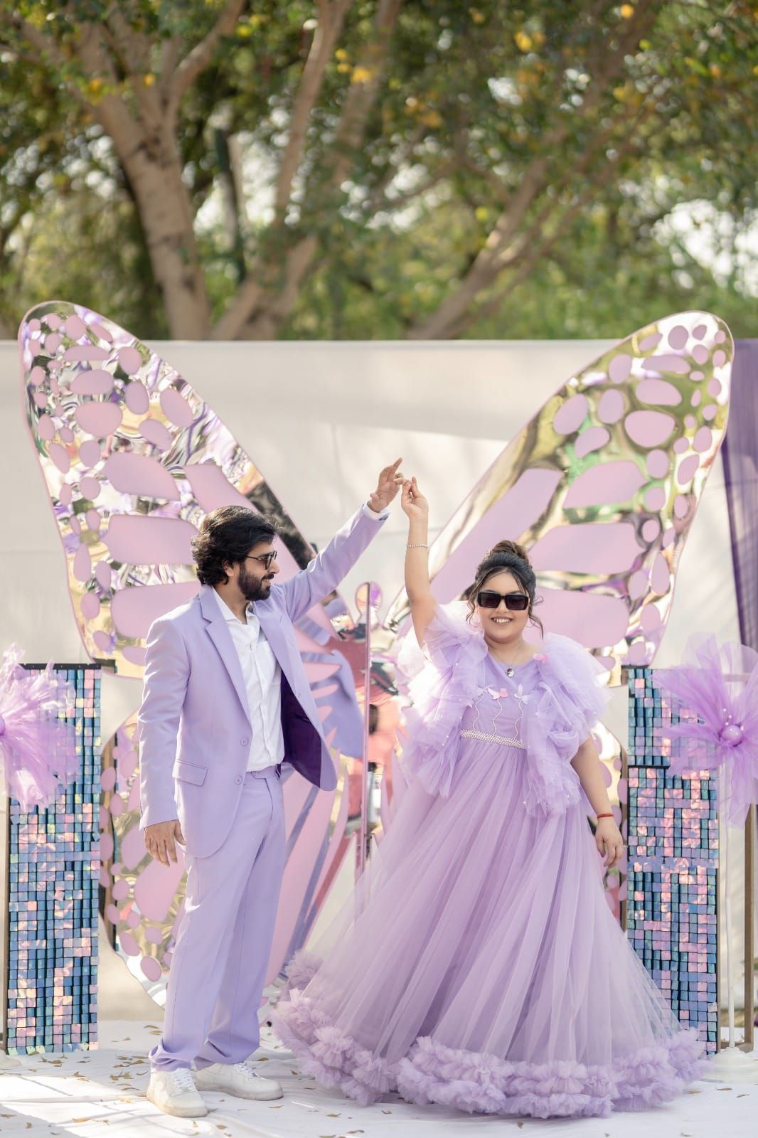 Couple in matching lavender outfits posing before a decorative butterfly backdrop at an outdoor celebration.