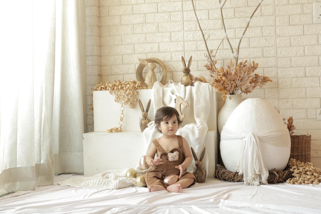 A toddler sits on a white bed surrounded by Easter decorations including bunny figurines and a large egg prop.
