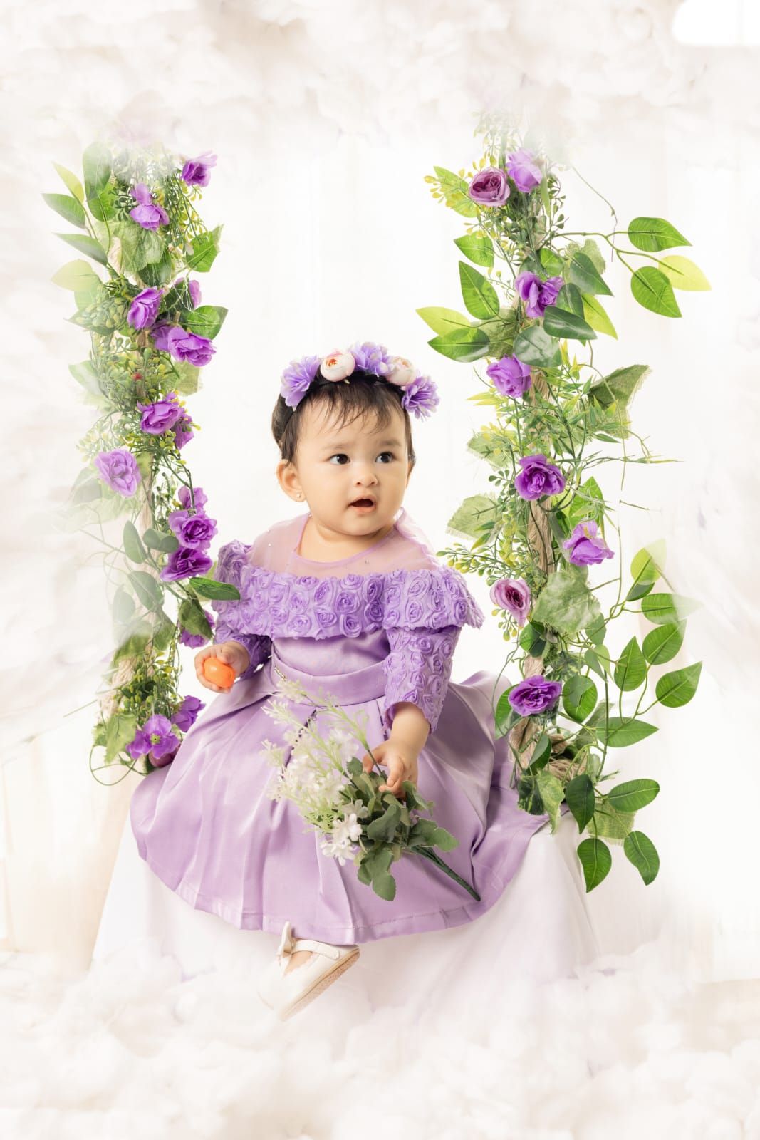 Baby girl in purple dress and floral crown sits on flower-adorned swing holding bouquet.