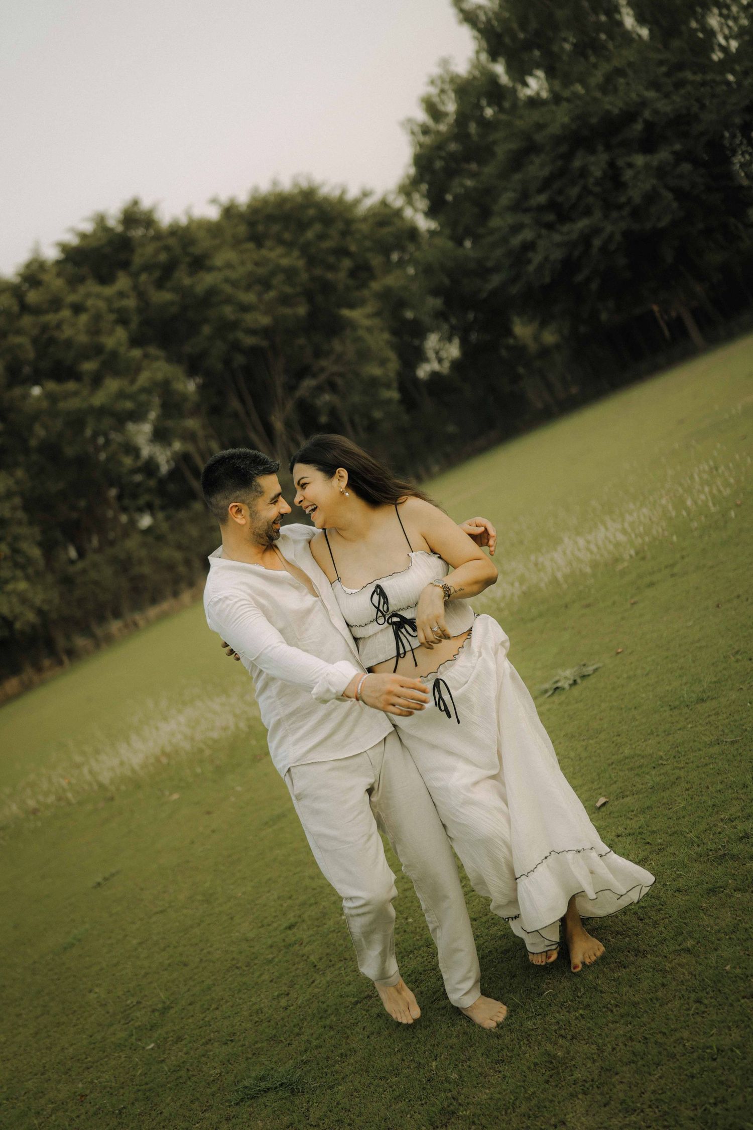 A couple in white outfits dance barefoot on green grass with trees in the background.